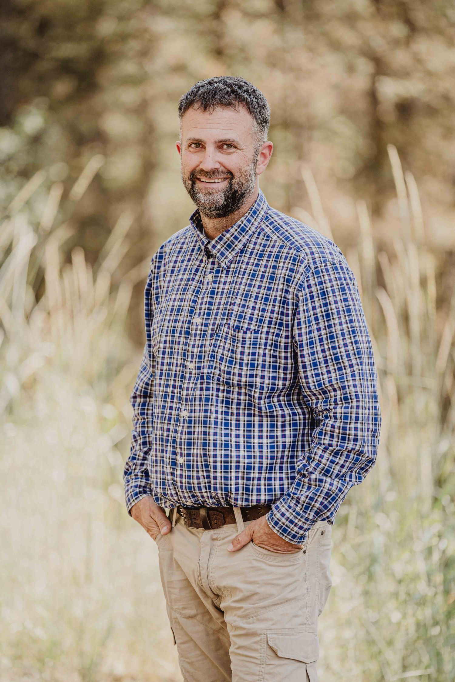 Person wearing blue plaid shirt and khaki pants poses outdoors in natural setting with blurred background.