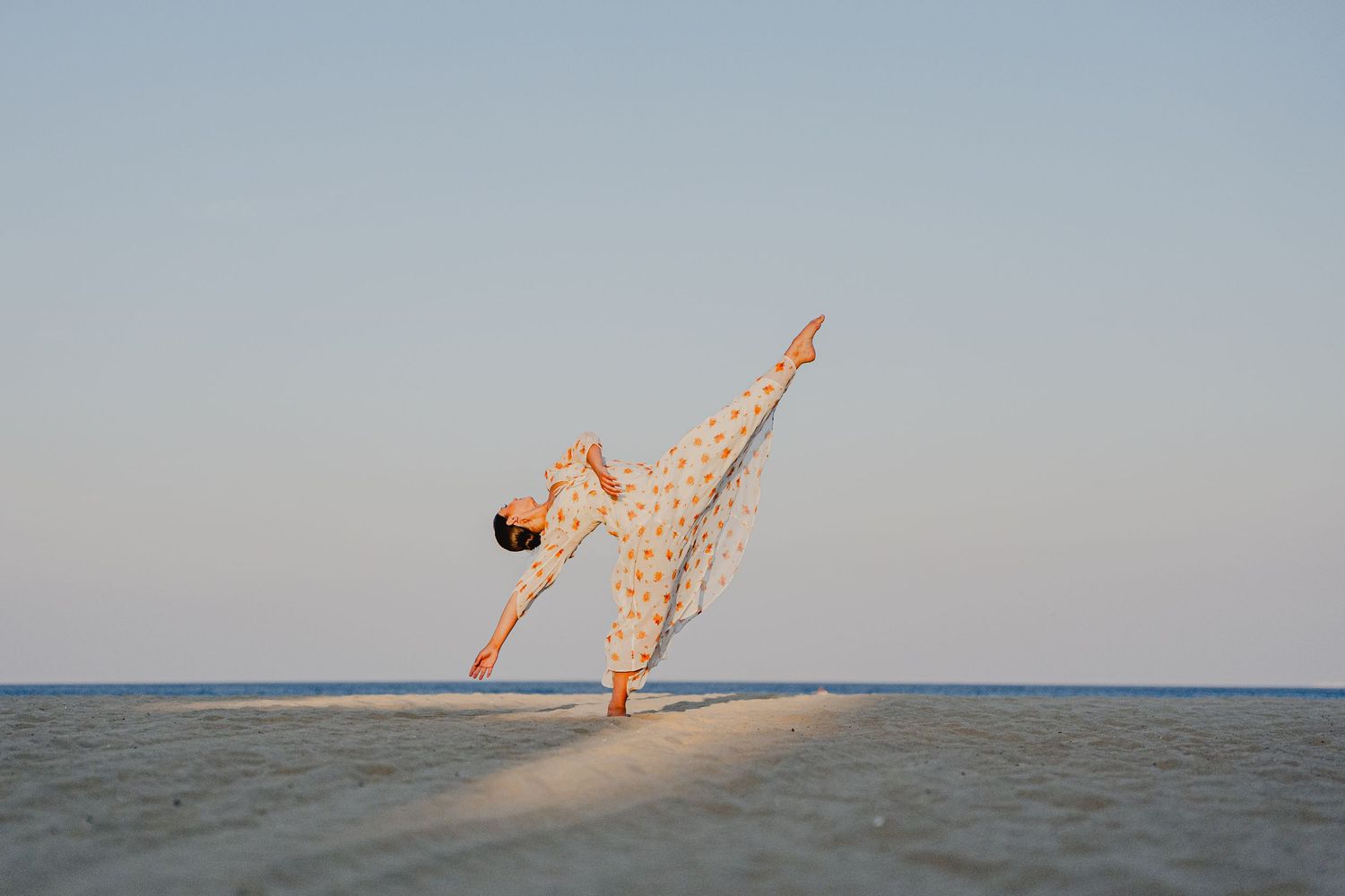 Woman in floral dress performing a graceful yoga pose on a sandy beach with calm blue sky.