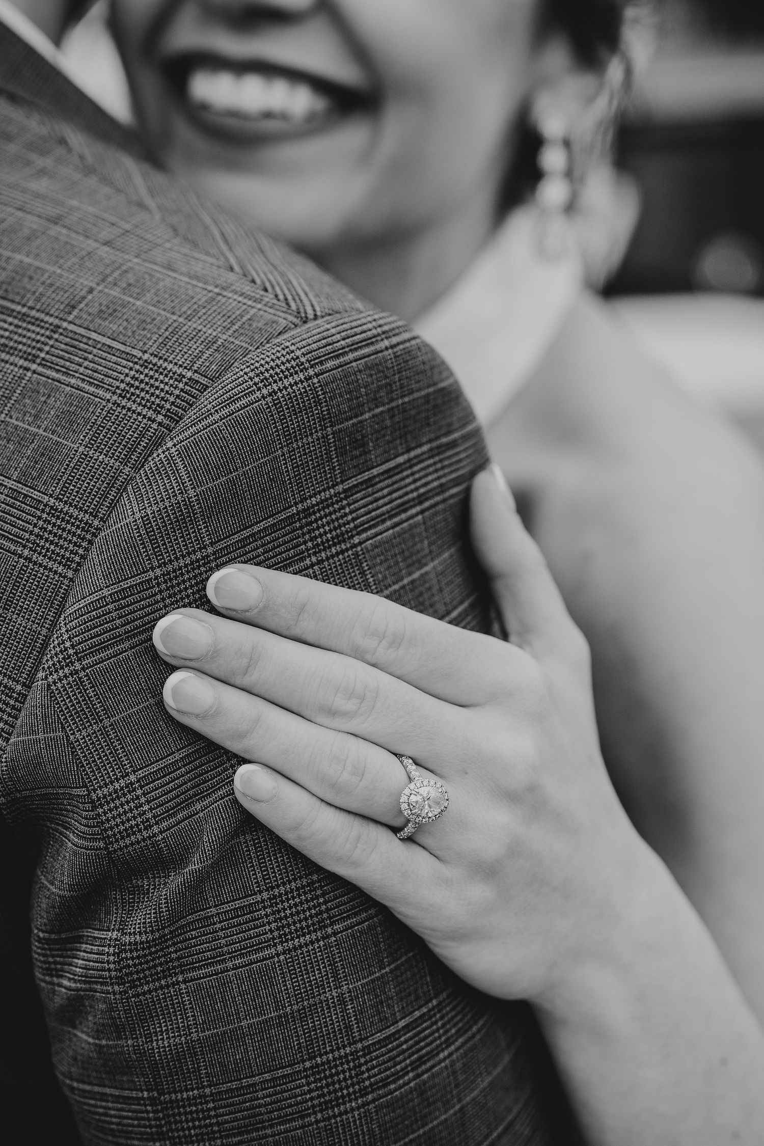 Black and white detail shot of a bride's hand with engagement ring resting on groom's plaid suit jacket.