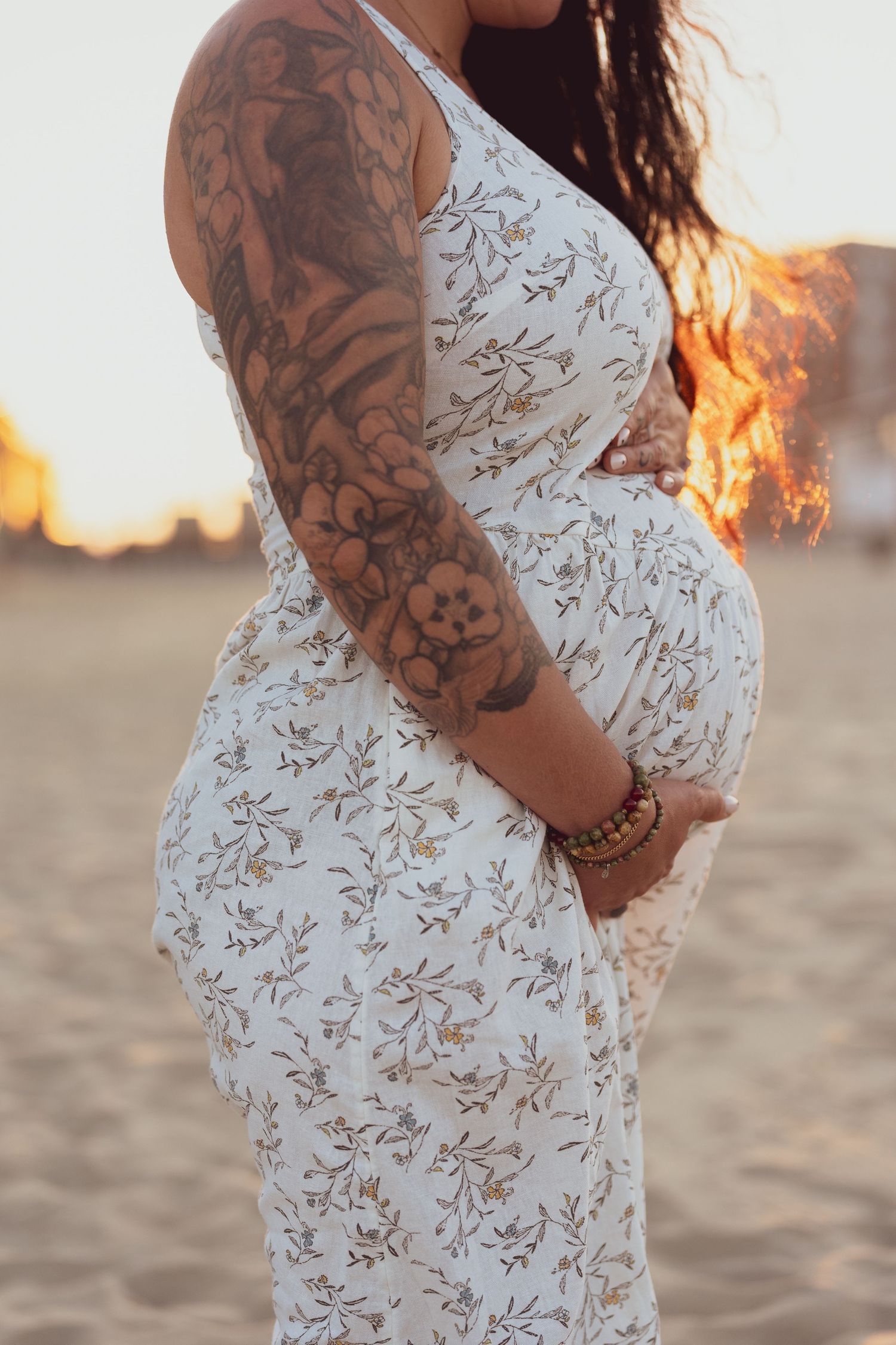 Close-up of a pregnant belly in a white floral dress with detailed tattoo sleeve.