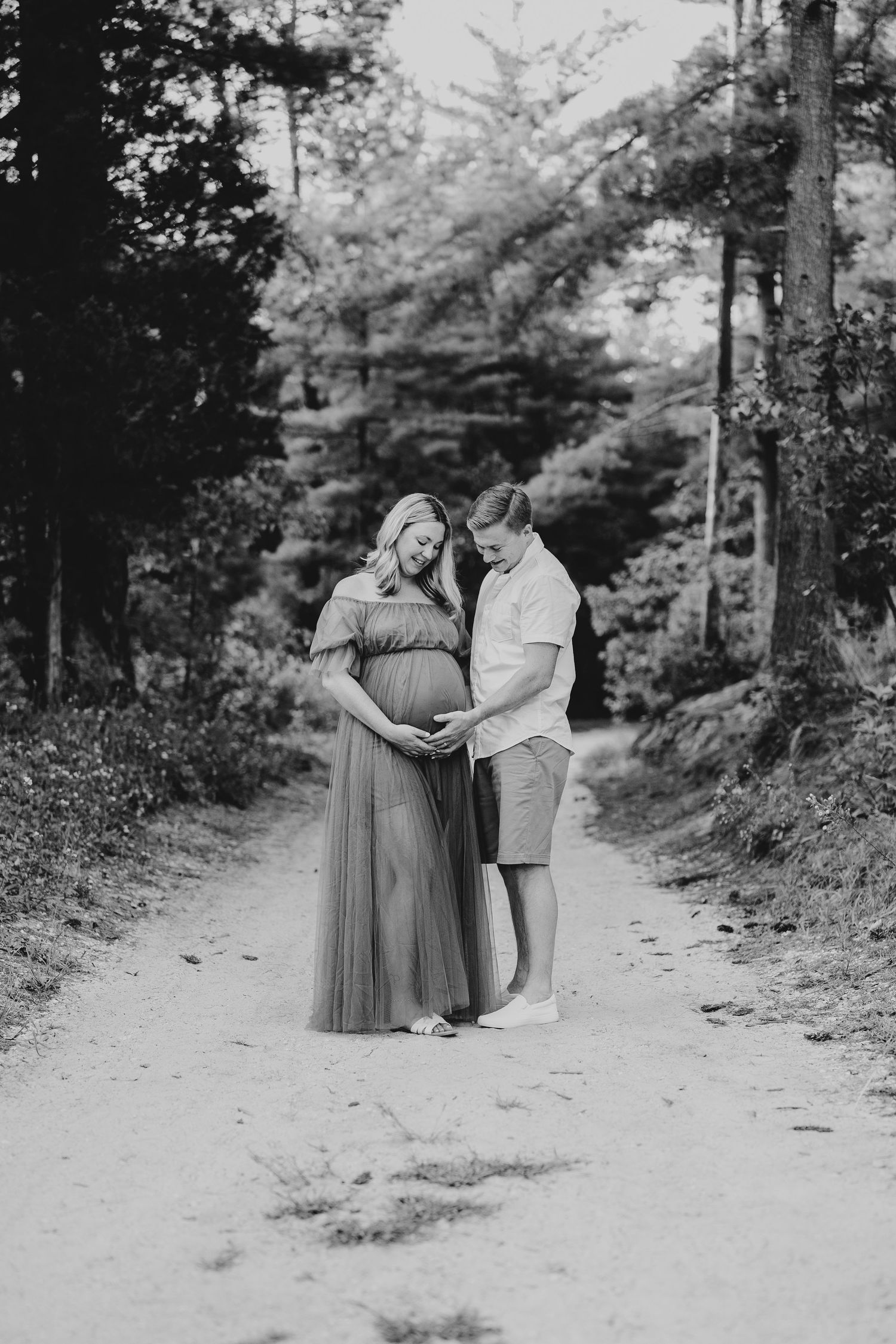 Black and white maternity photo of a couple embracing on a forest path.