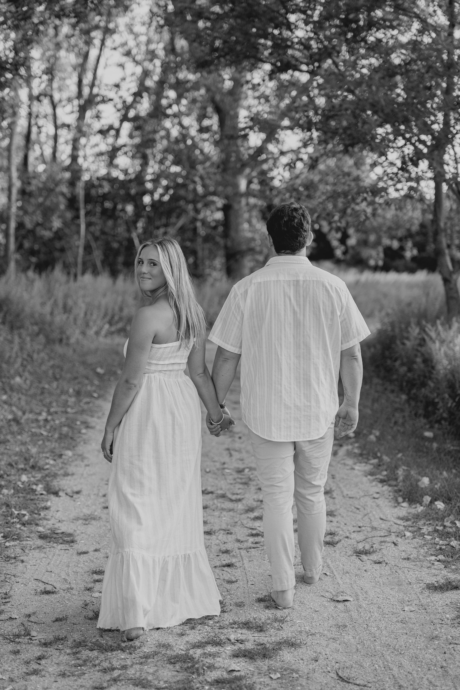 Black and white photo of a couple holding hands walking on a dirt path through trees, woman in white dress looking back.