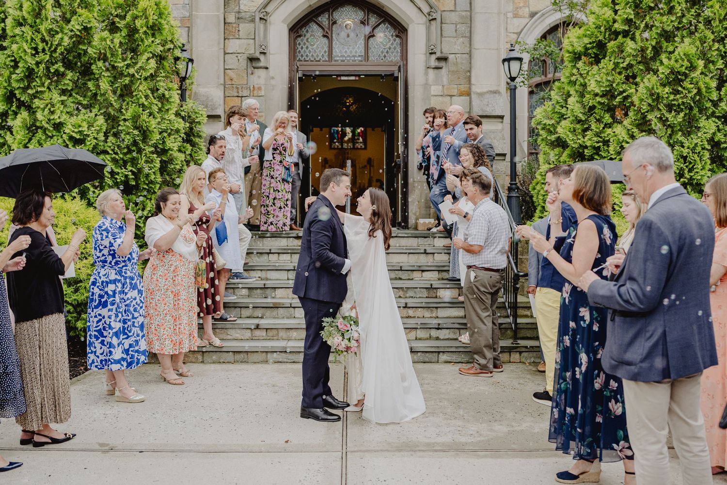 Newlyweds share a kiss outside a stone church as wedding guests line the steps cheering in celebration.