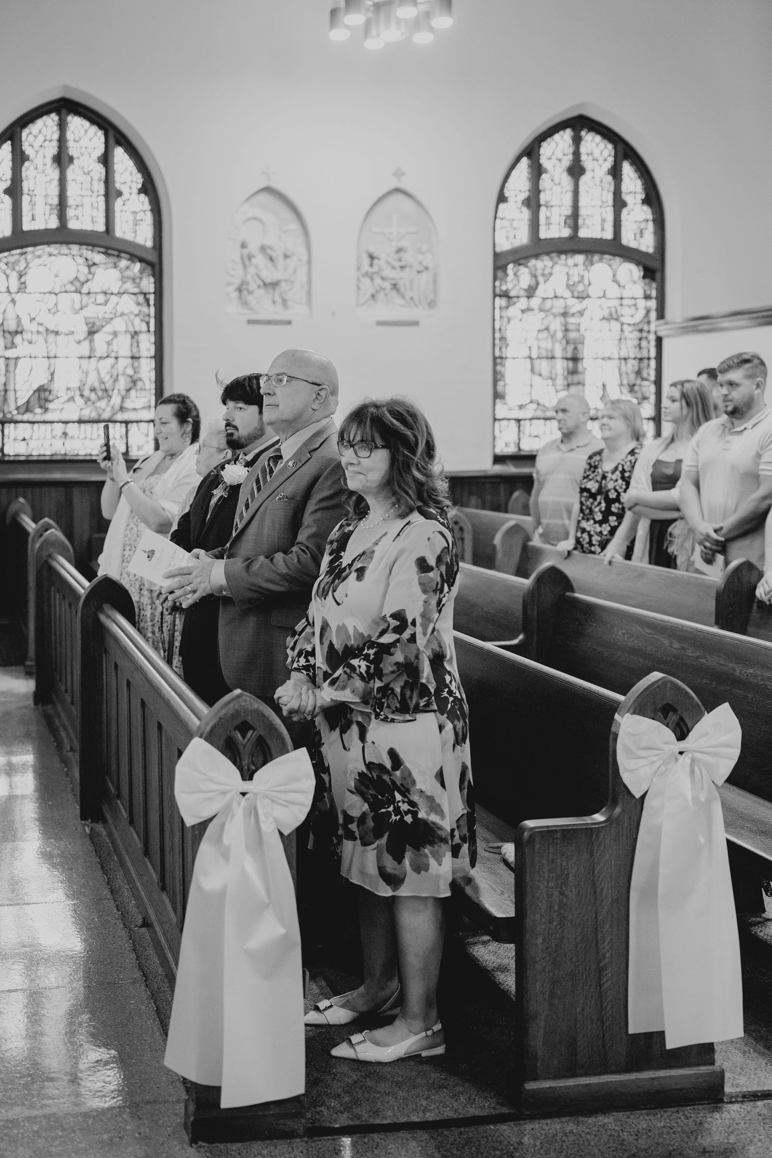 Black and white photo of wedding guests standing in church pews adorned with white ribbon bows during a ceremony.
