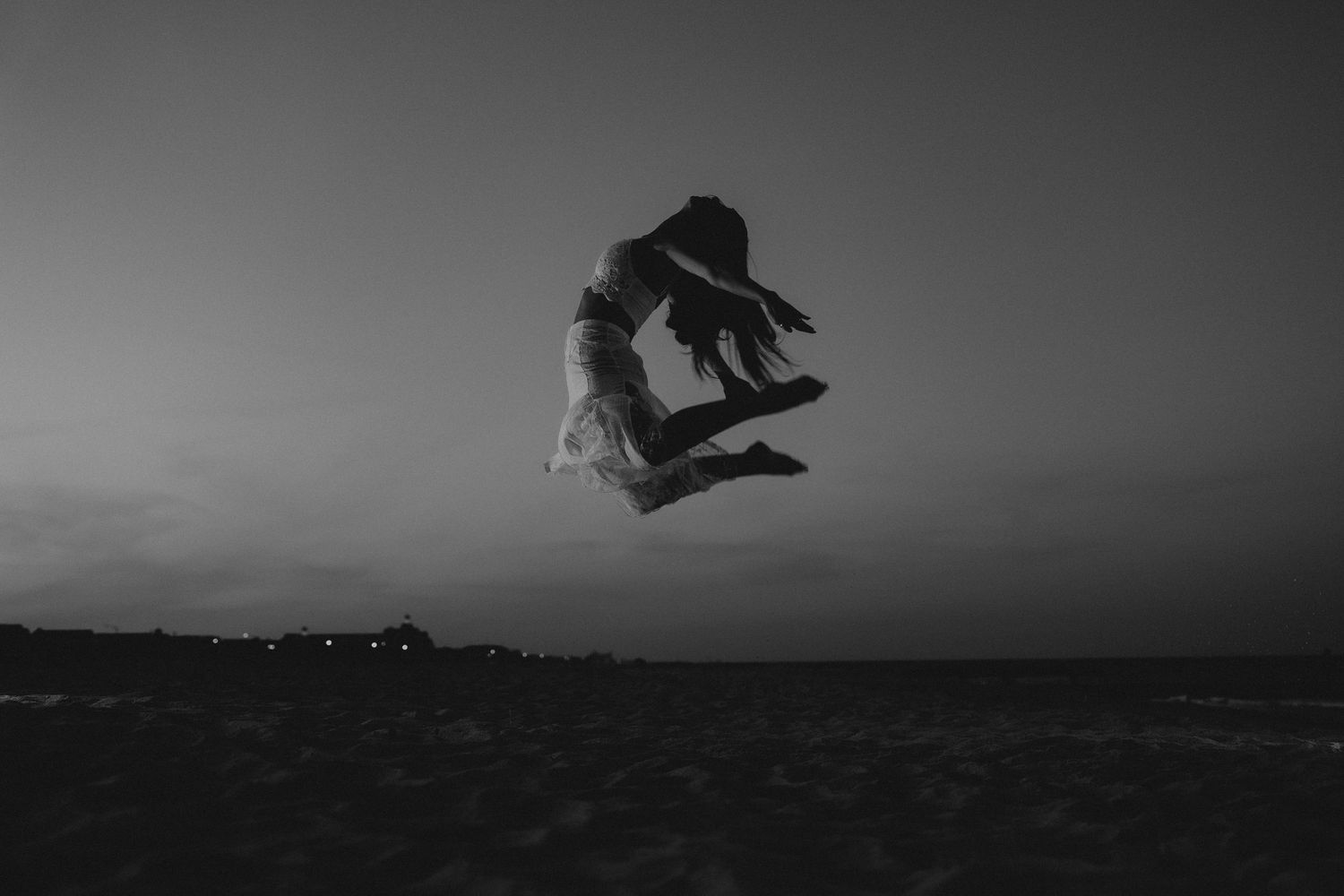 Black and white photo of a woman leaping gracefully against a twilight sky on a beach.