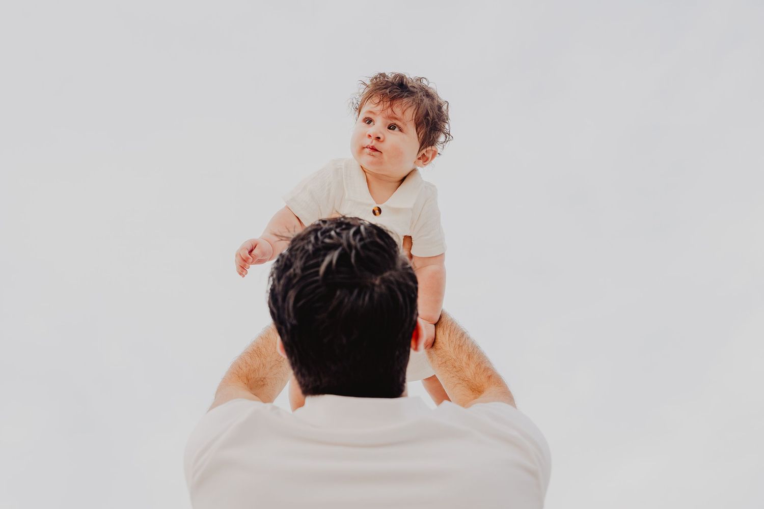 A father lovingly lifts his curly-haired baby up in the air against a white background.