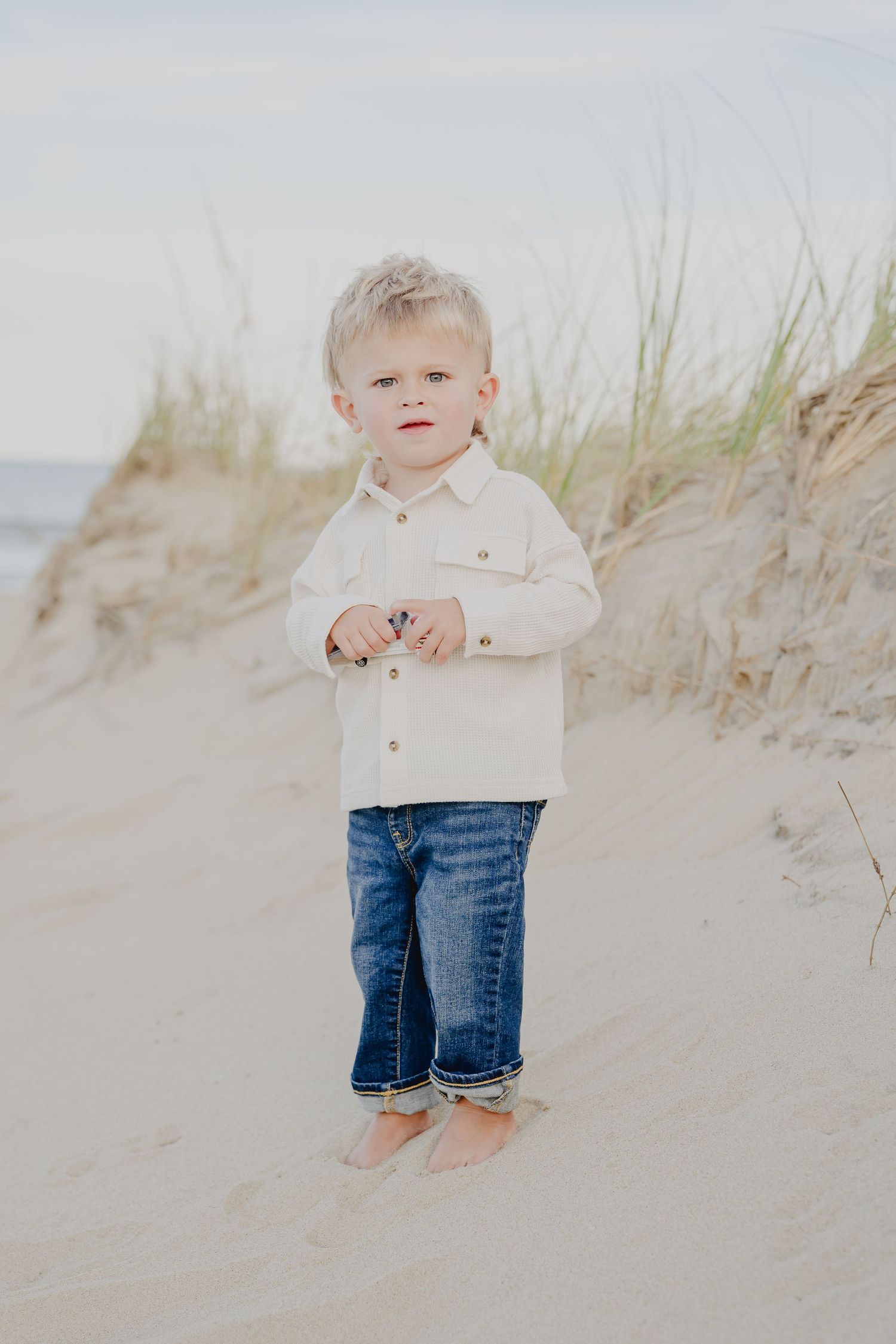 Toddler boy in white shirt and jeans standing barefoot on sandy beach dunes.