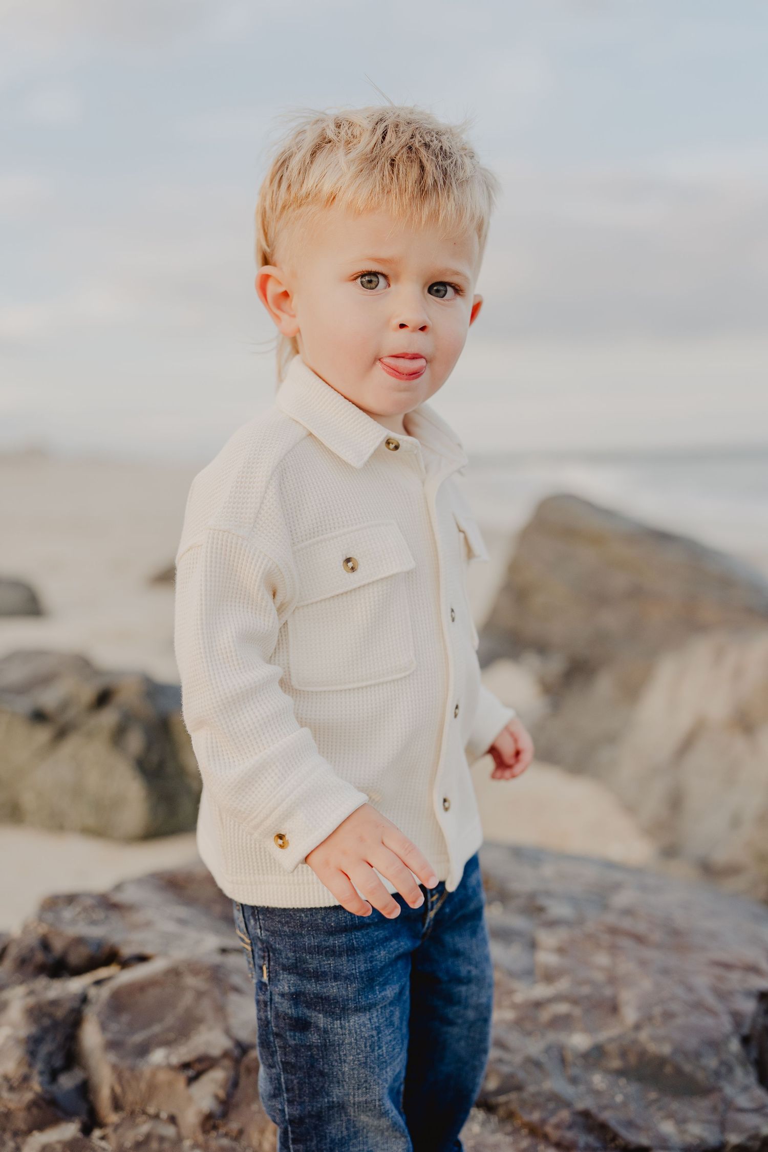 Blonde toddler boy in white shirt and jeans sticking out tongue at rocky beach.