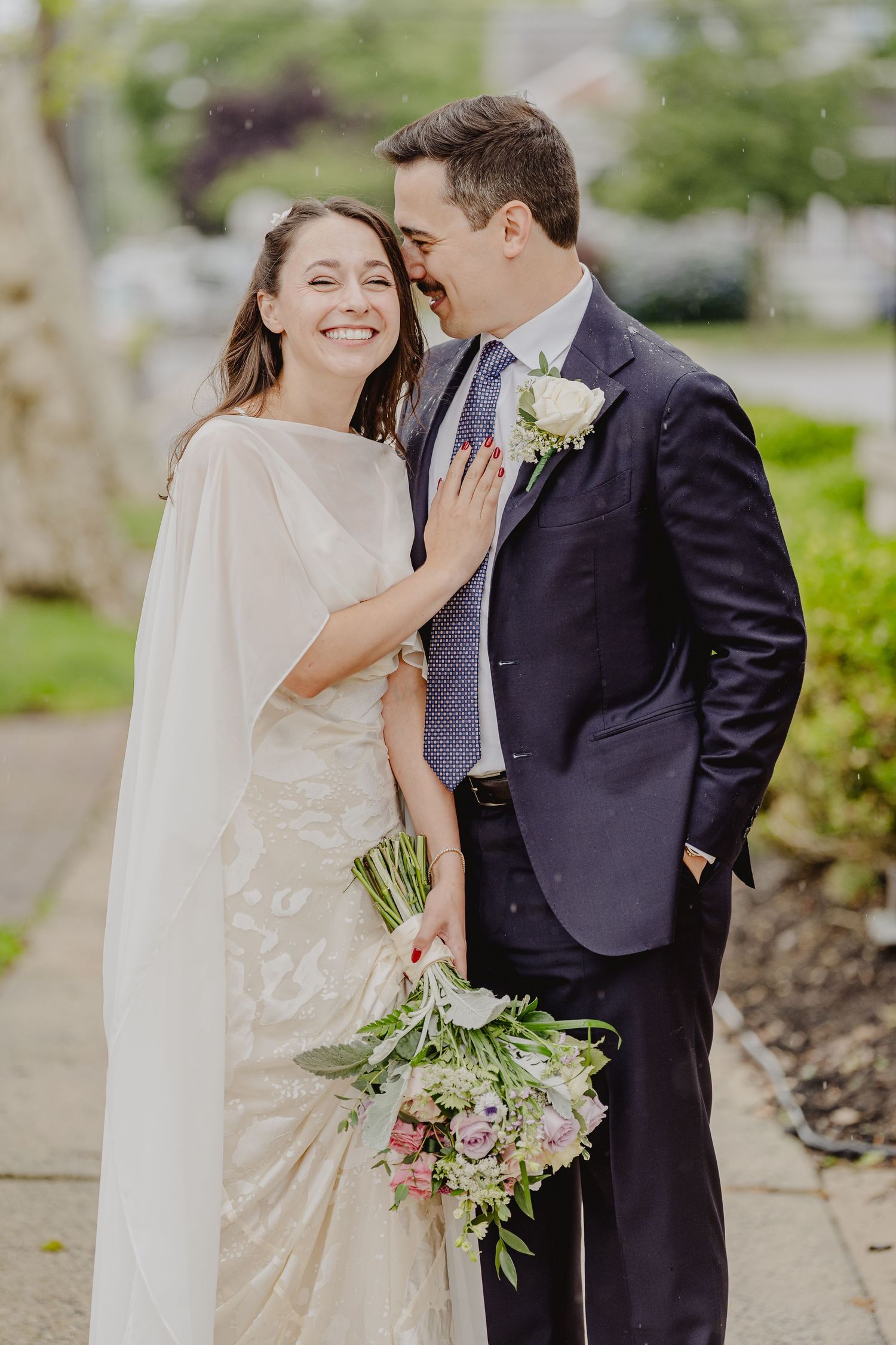 Smiling bride in white cape gown holds floral bouquet beside groom in navy suit with rose boutonniere outdoors.