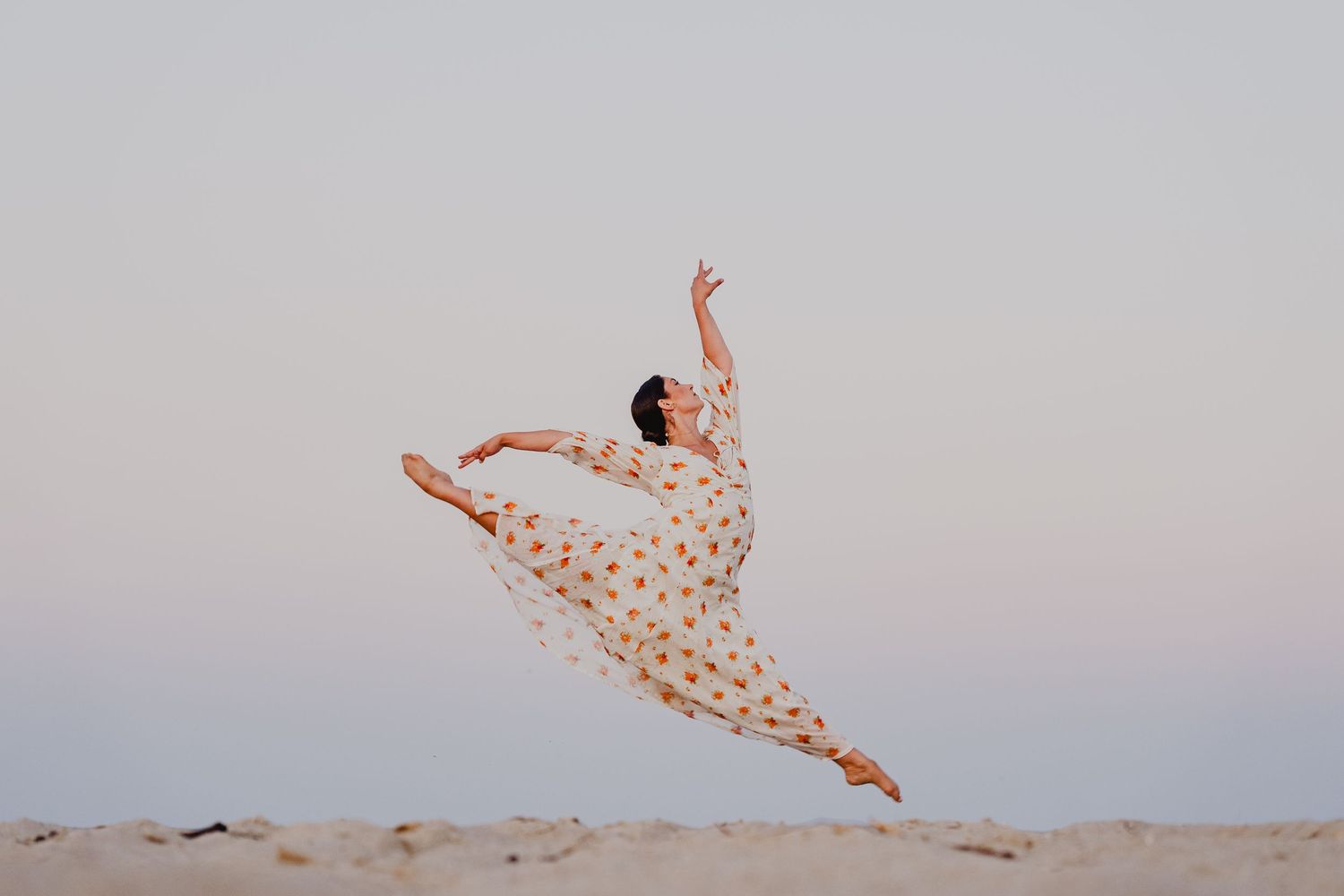 Graceful ballerina leaping mid-air on sandy beach in floral dress against soft twilight sky.