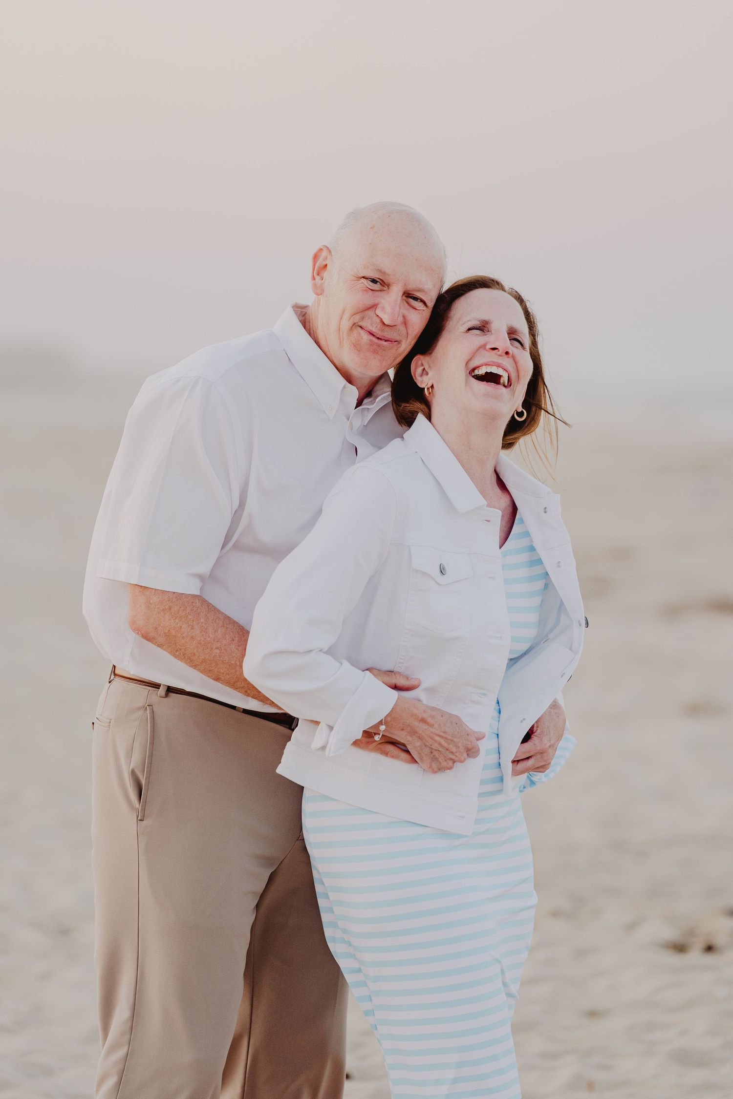 Happy elderly couple embracing and laughing together on a sandy beach, wearing casual white outfits.