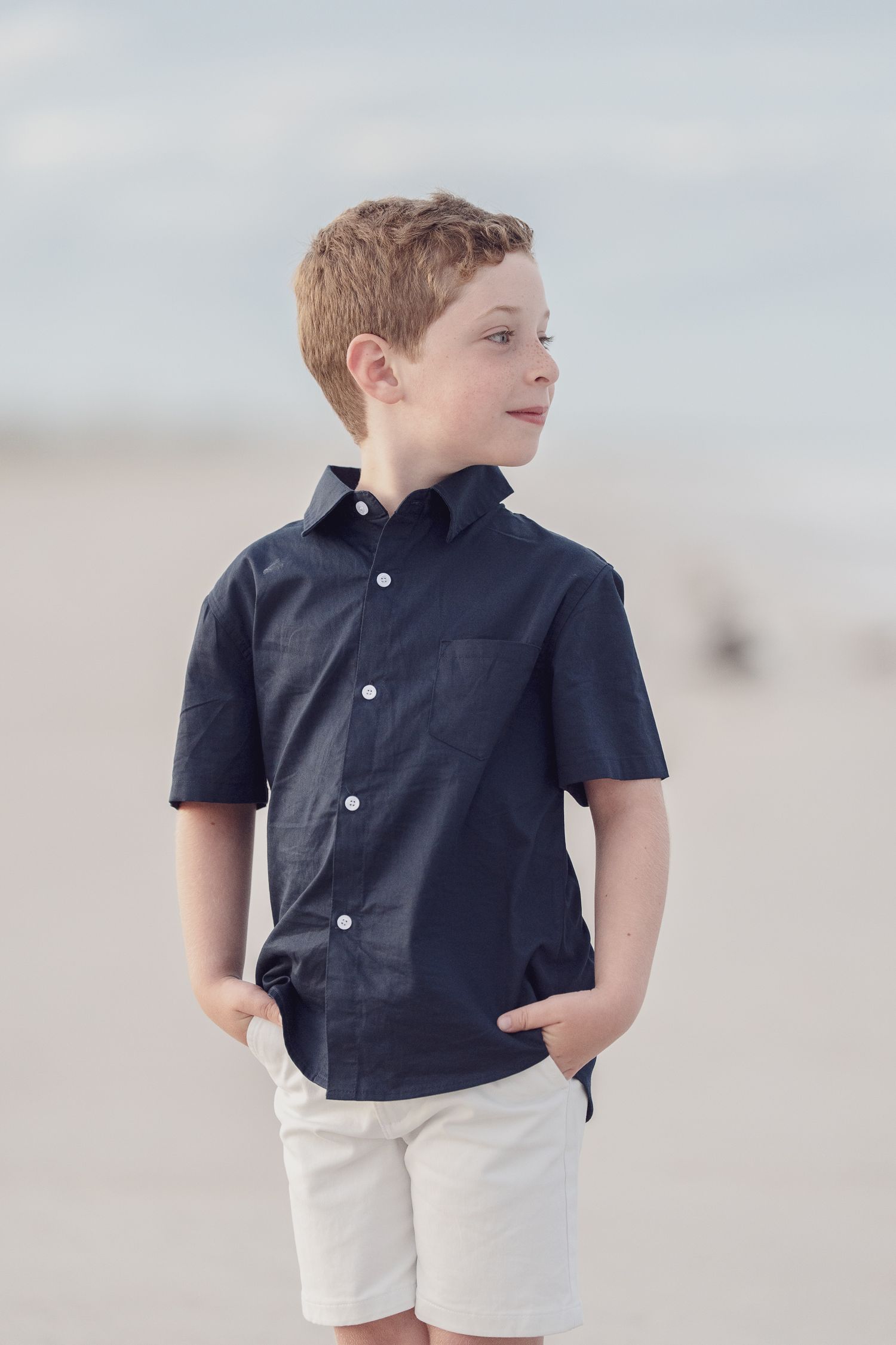 Child in dark polo and white shorts posing casually on beach.