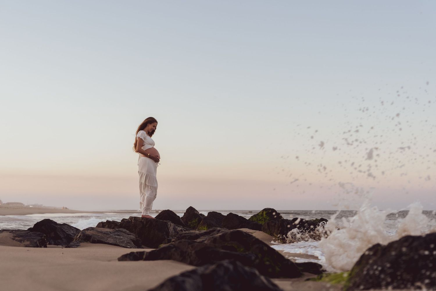 Ocean waves crash against rocky shore as figures stand on beach at sunset.