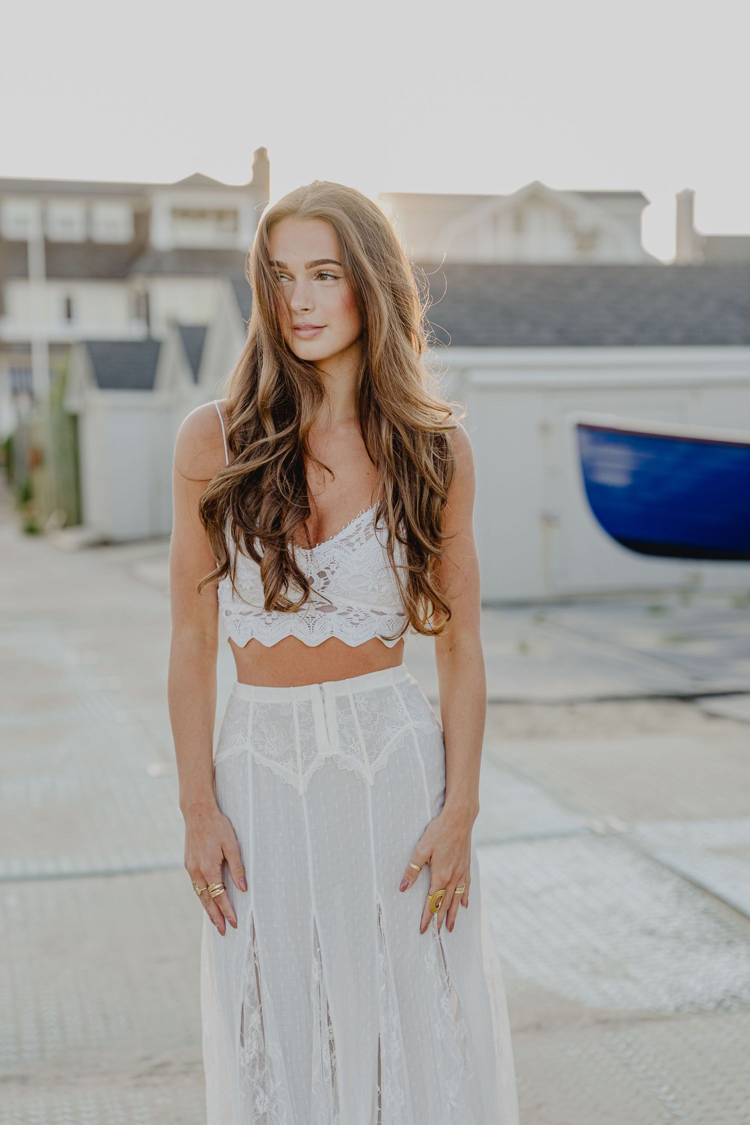 Young woman in white lace crop top and sheer maxi skirt posing outdoors near coastal buildings in golden hour light.