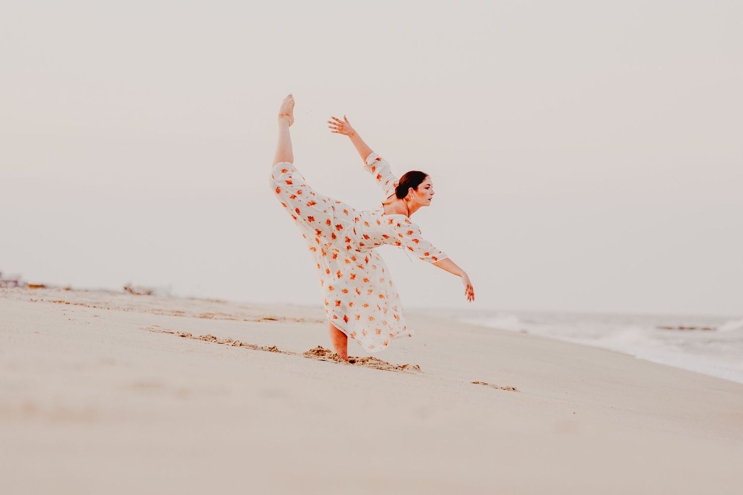 A graceful dancer performs an elegant arabesque on a sandy beach in a floral dress at golden hour.