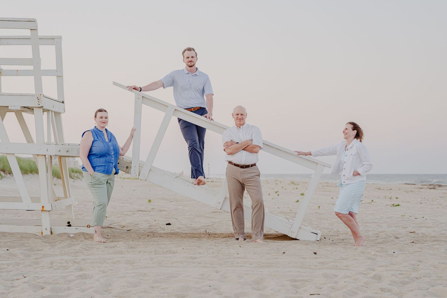 Family of four posing playfully on sandy beach near lifeguard stand, holding white fabric, smiling outdoors.