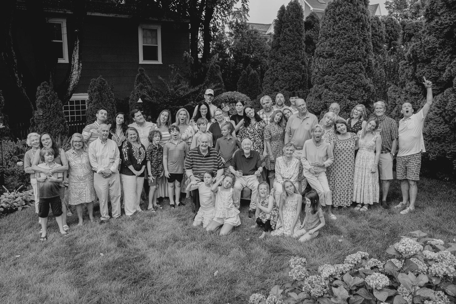 Large family group portrait outdoors in black and white, posing together on a lawn with trees and a brick house behind them.