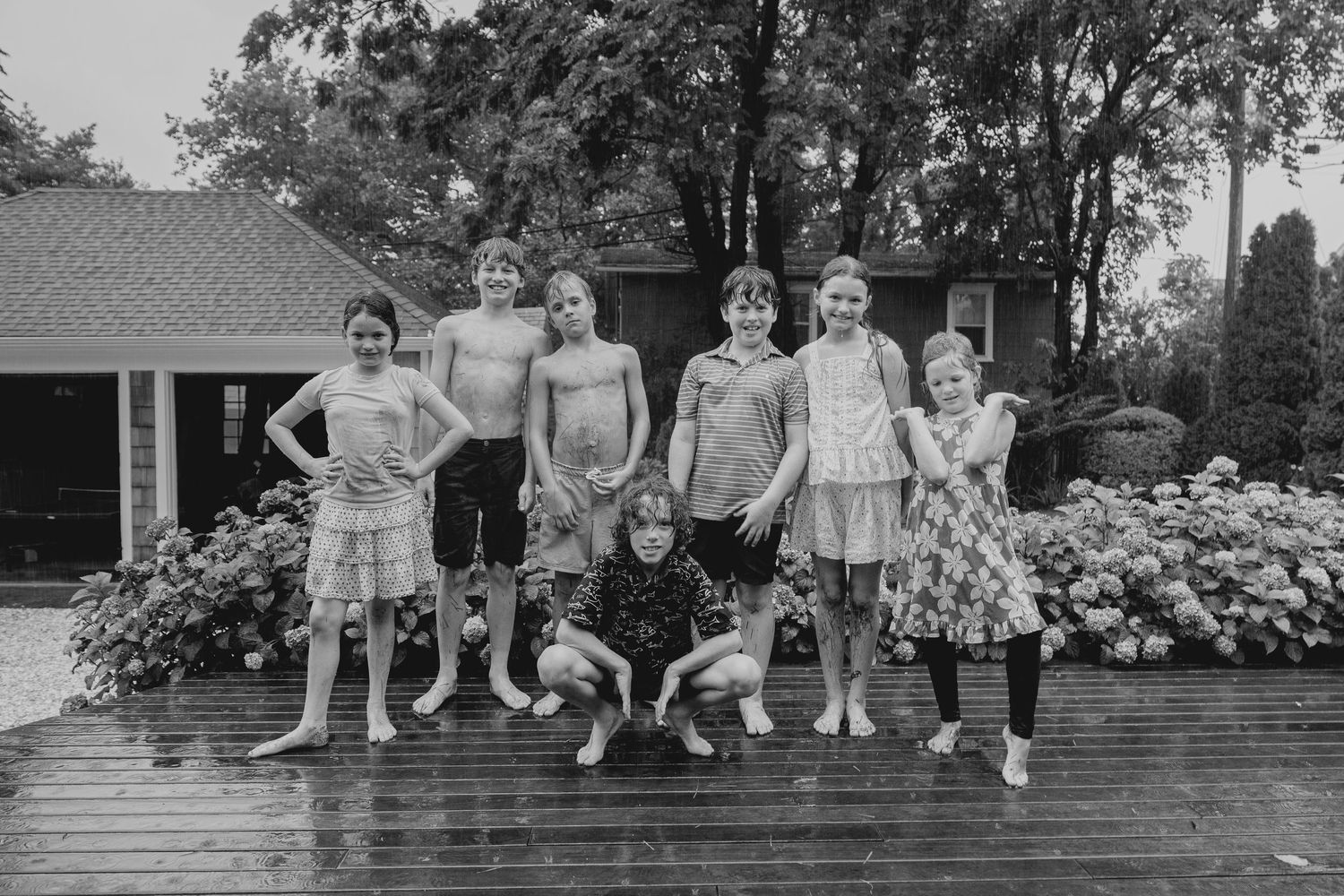 Black and white photo of a group of children posing together on a wet deck outside a house.