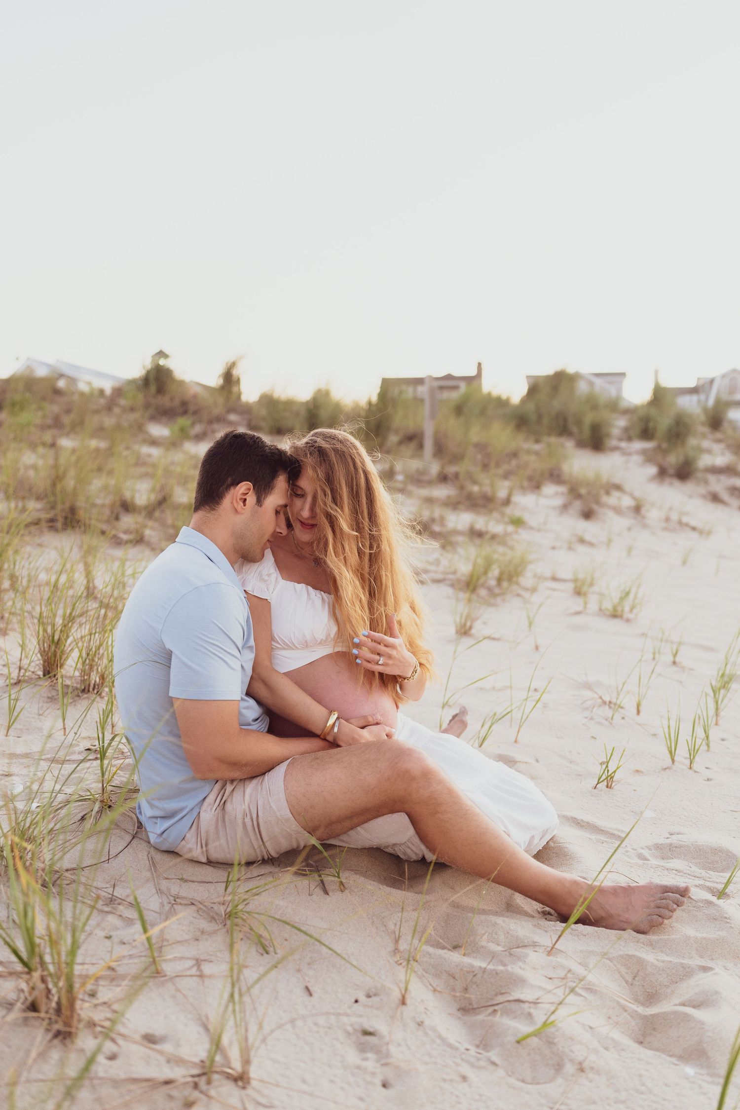 A couple sits intimately together on beach sand surrounded by dune grass.