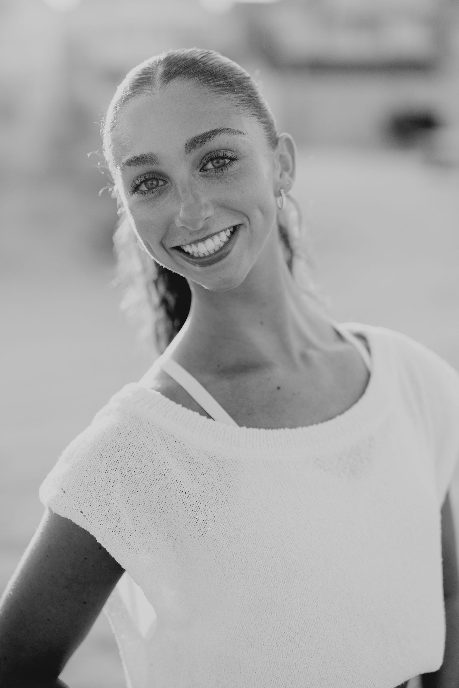Black and white portrait of a smiling young woman with braided hair wearing a white top outdoors.