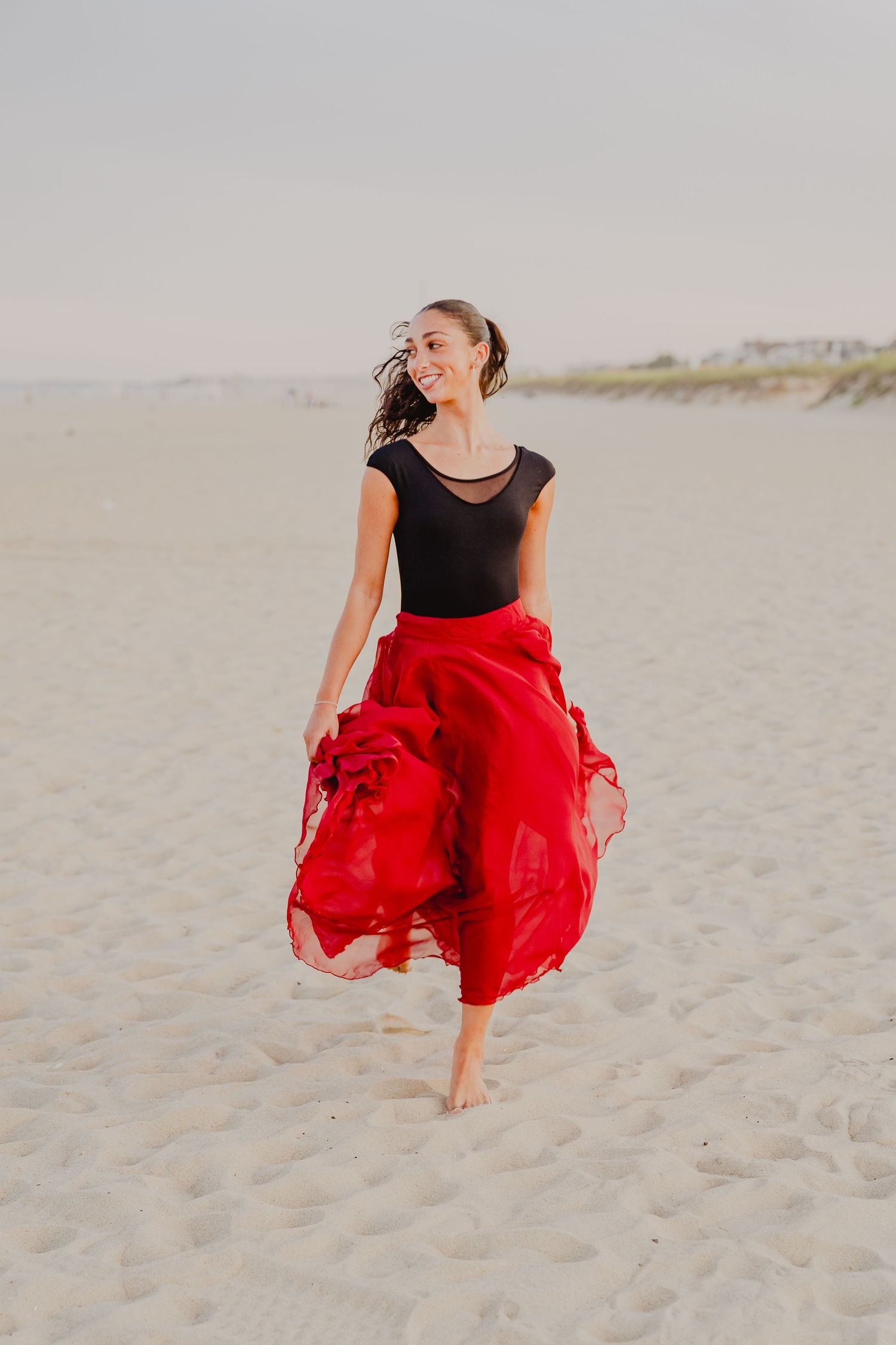 Smiling dancer in vibrant red flowing skirt and black top twirls barefoot on a sandy beach.
