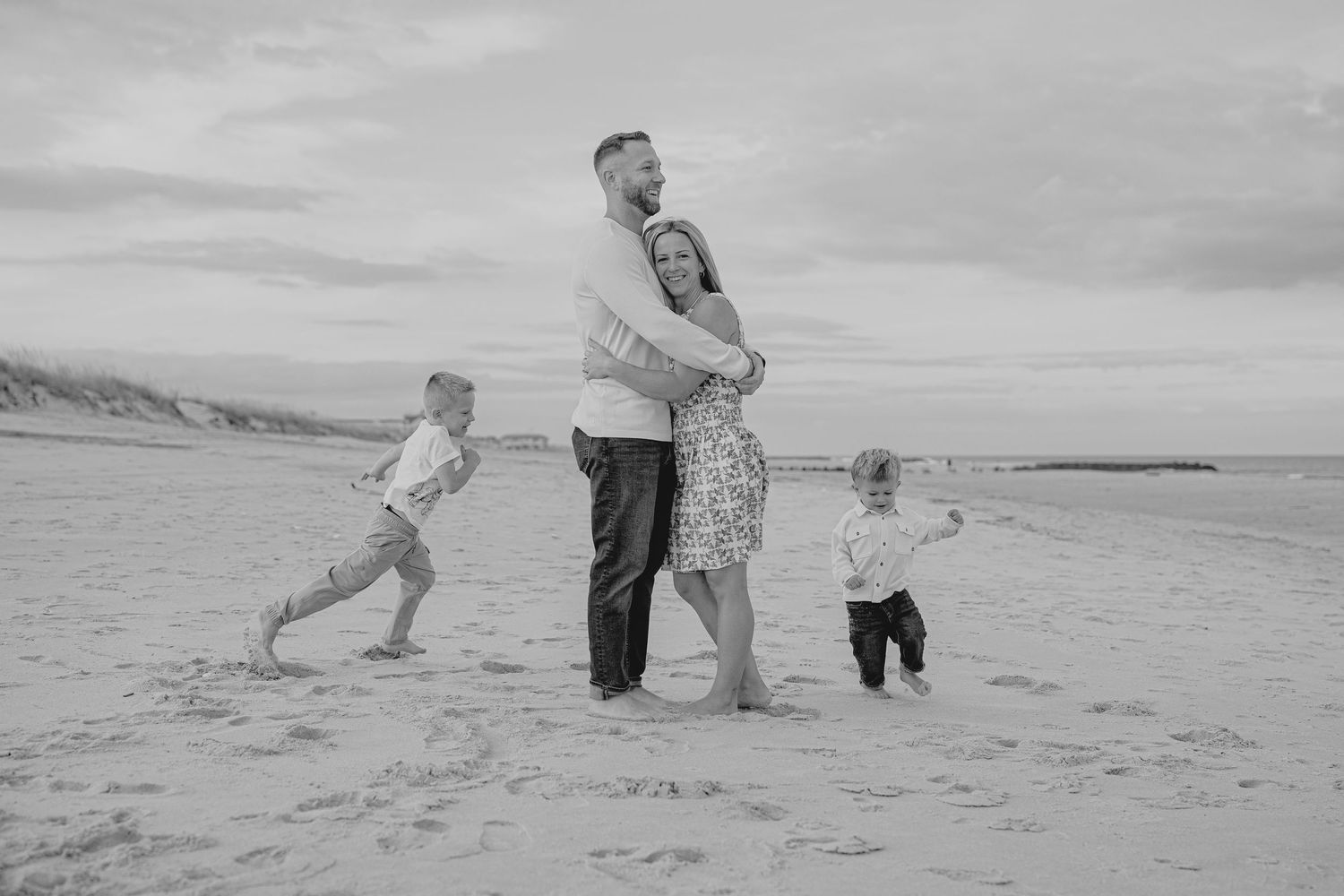 Black and white beach family photo with couple embracing and two young children playing on sandy shore.
