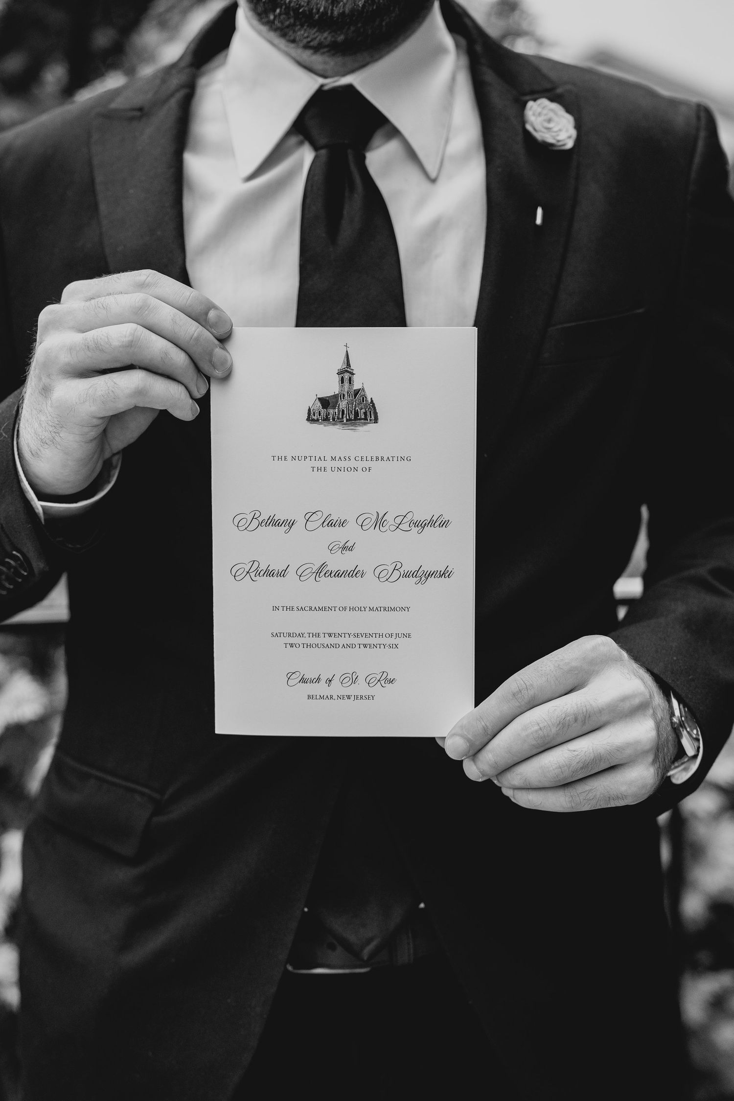 Groom in black suit holds wedding ceremony program card at church in elegant black and white photo.