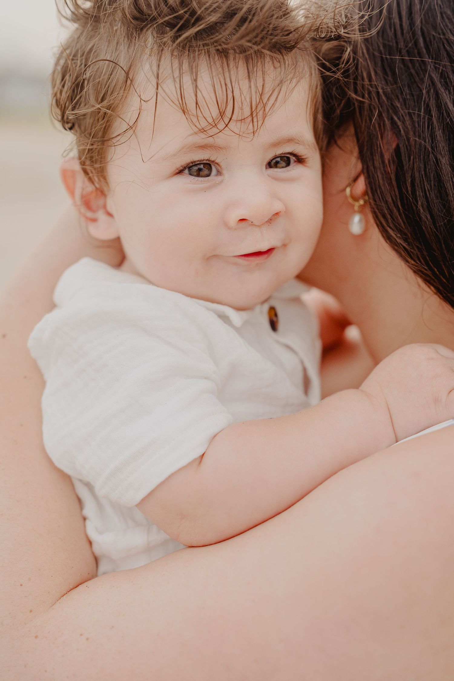 A smiling baby with wet hair in a white outfit being held by a woman wearing a pearl earring outdoors.