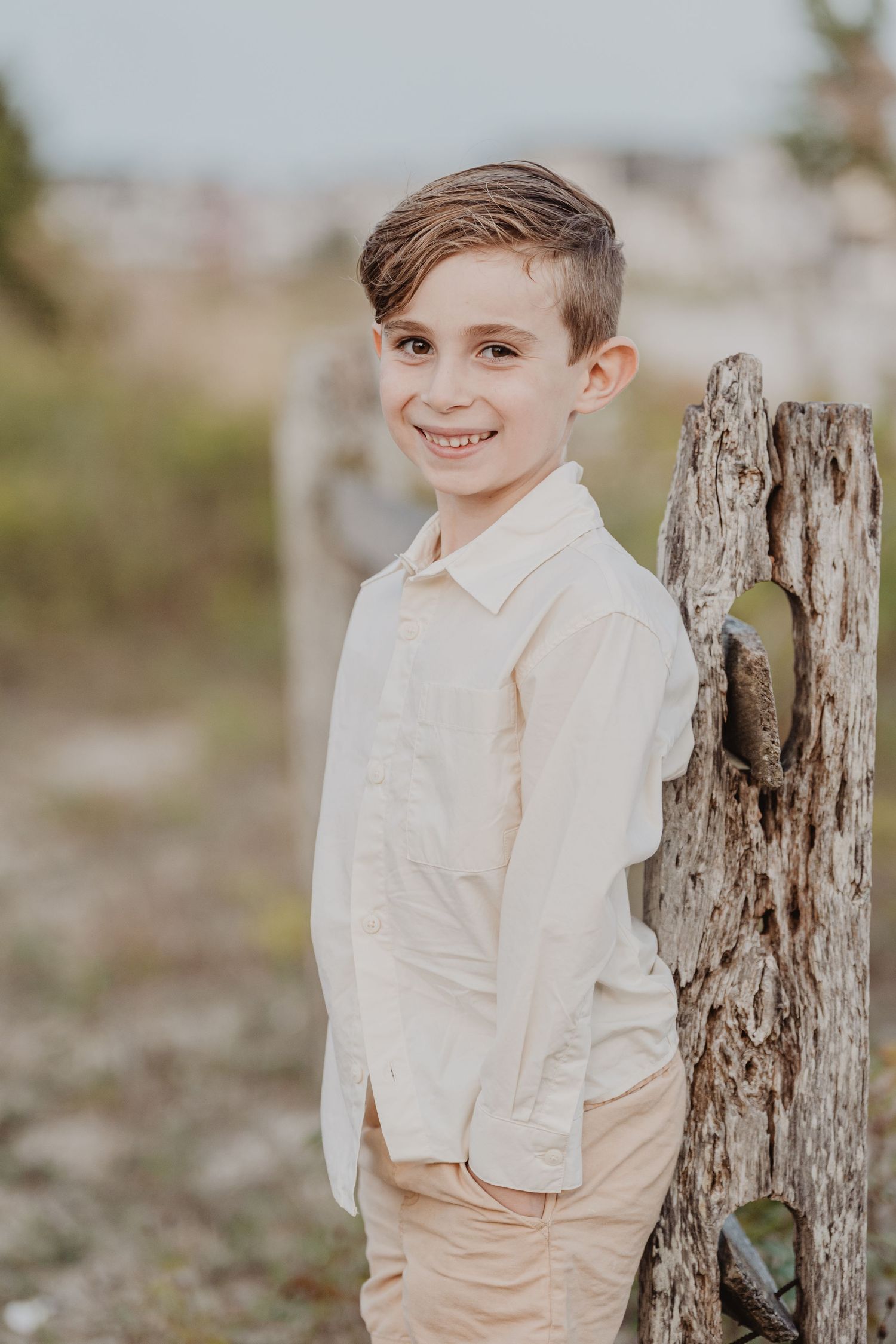 Young boy in white dress shirt poses casually against rustic wooden fence post.