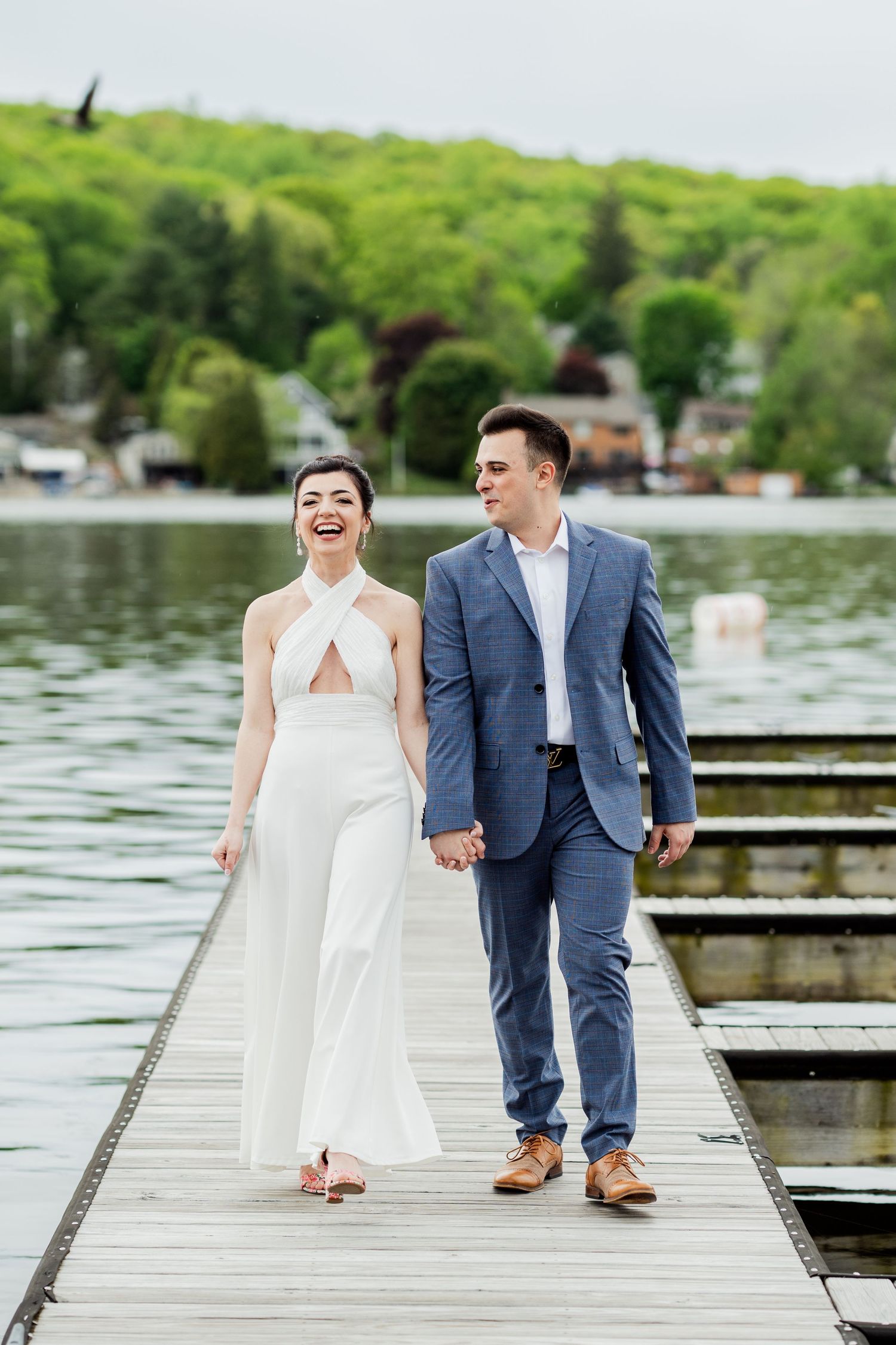 Newlyweds walk hand in hand on a lakeside dock surrounded by green trees.