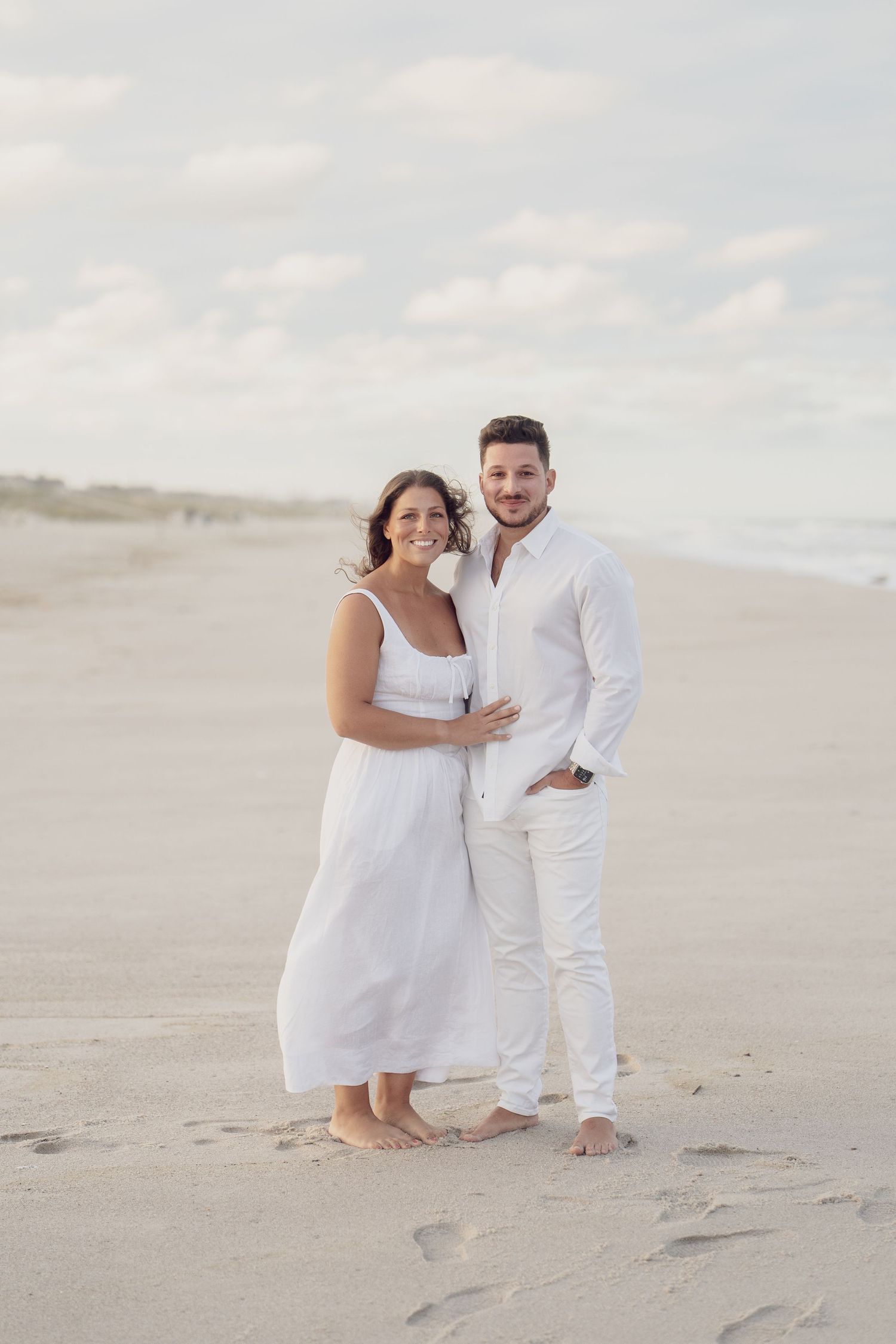Romantic beach photoshoot featuring couple in coordinated white outfits against soft clouds.