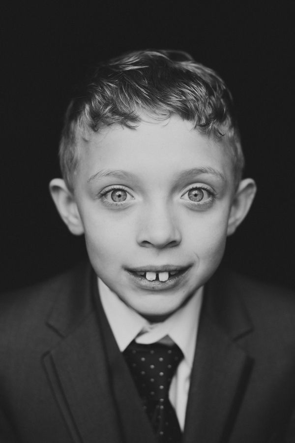 Black and white portrait of a young boy in a suit and tie, smiling with gap teeth against dark background.