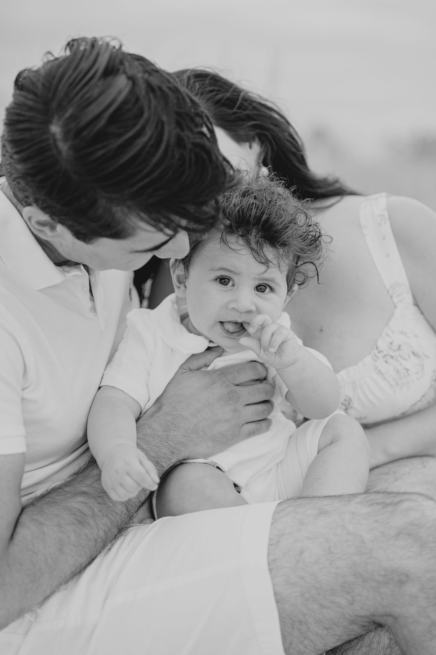 Black and white photo of parents lovingly holding their curly-haired baby, sharing a tender family moment outdoors.