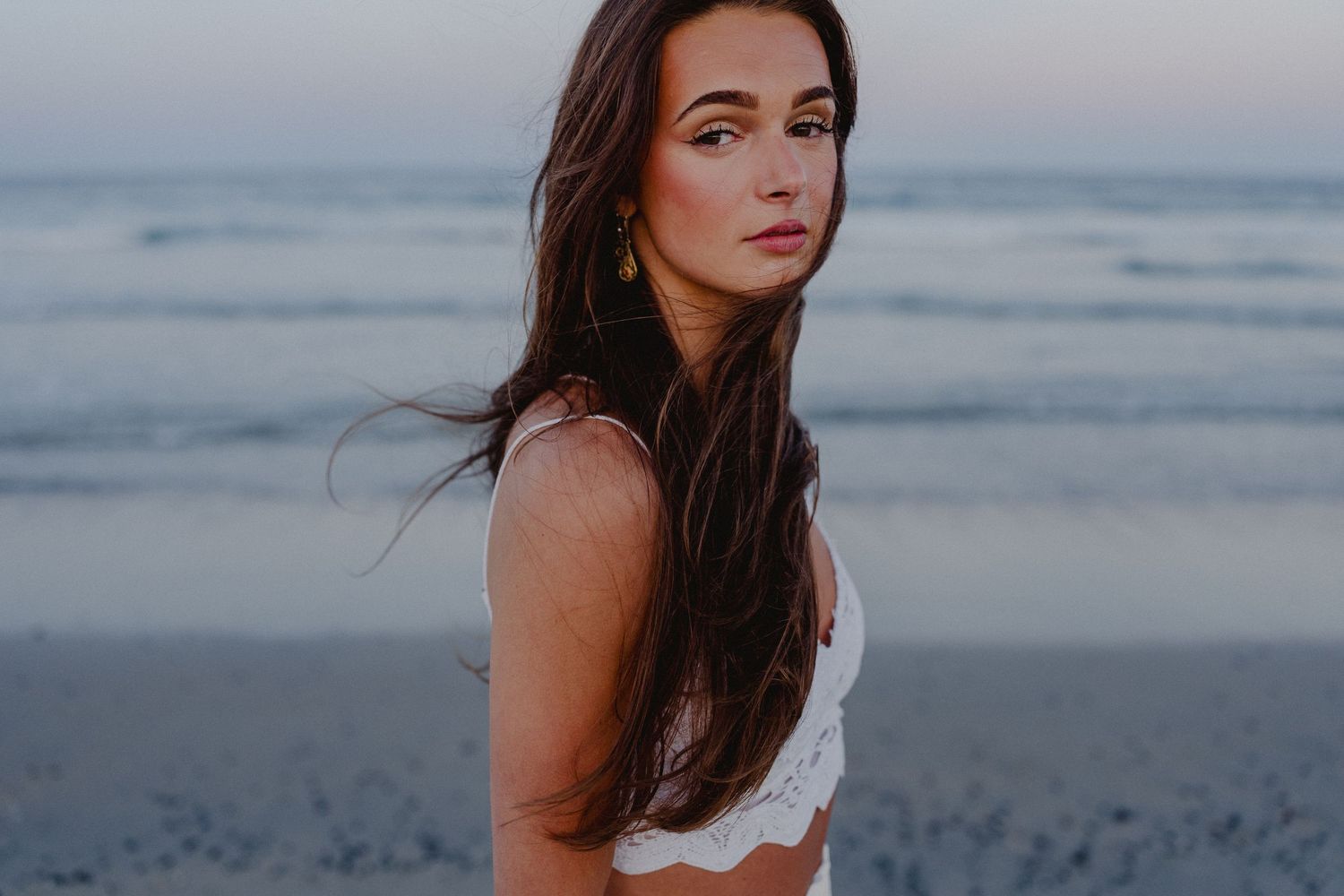 Young woman with long brown hair posing on a beach at dusk wearing a white crop top.
