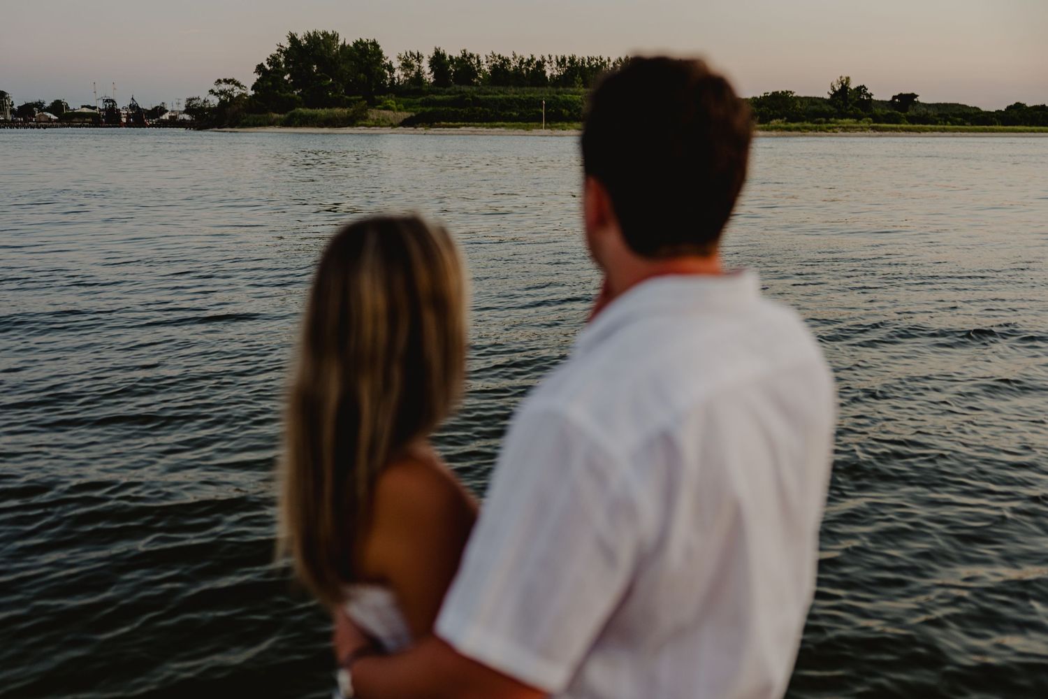 Couple embracing while gazing at a serene lake with lush green trees at sunset.