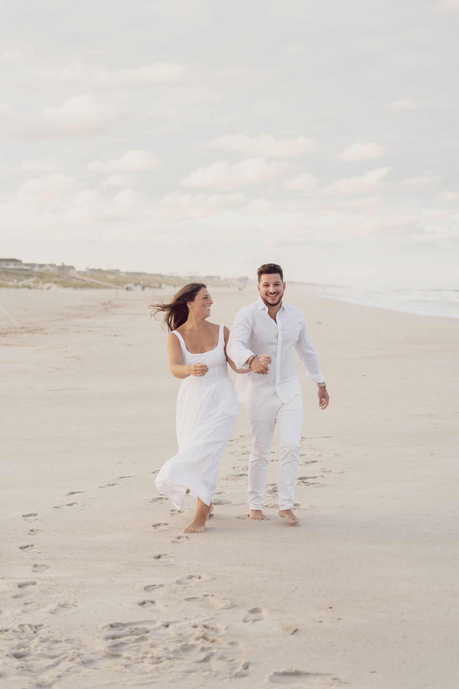 A couple in white outfits runs playfully along a sandy beach during a romantic photoshoot.