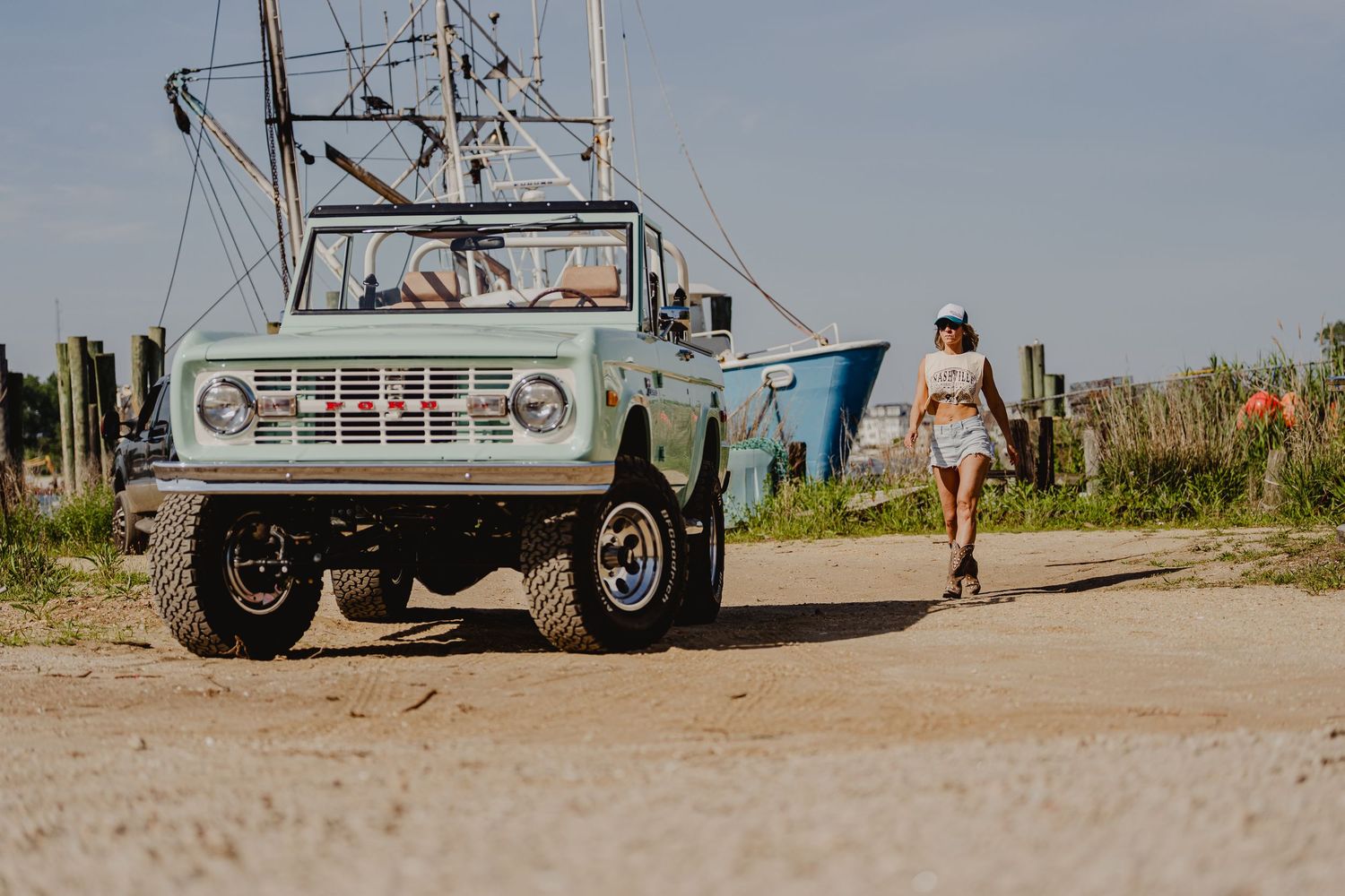 Vintage mint Ford Bronco parked on sandy path near shrimp boat with woman walking away in background.