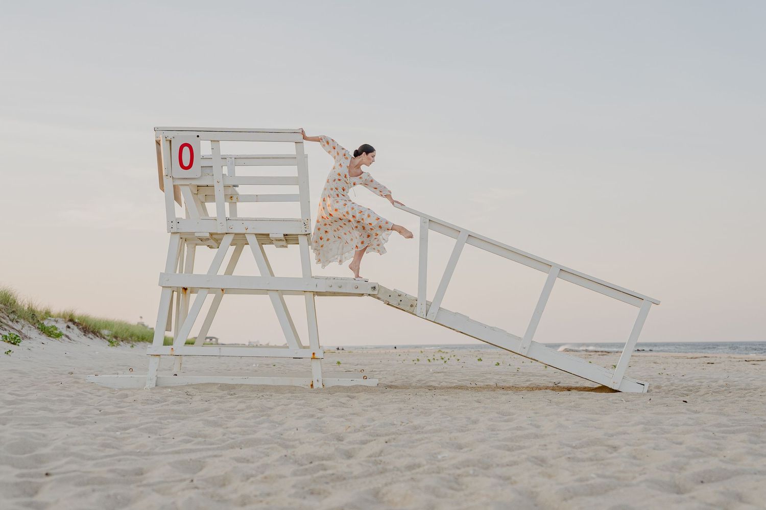 Woman in floral dress jumping off a white lifeguard stand on a sandy beach at sunset.