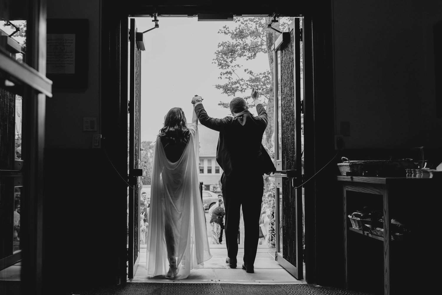 Black and white photo of bride and groom holding hands, walking through open doors at their wedding.