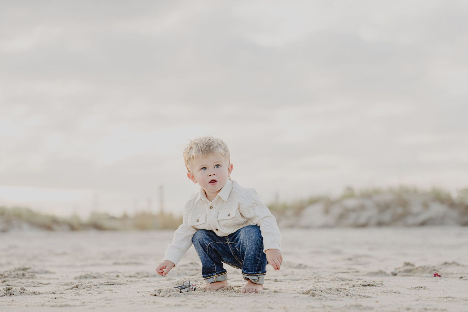 A toddler boy in jeans and white shirt crouches on sandy beach, exploring the shore on a cloudy day.
