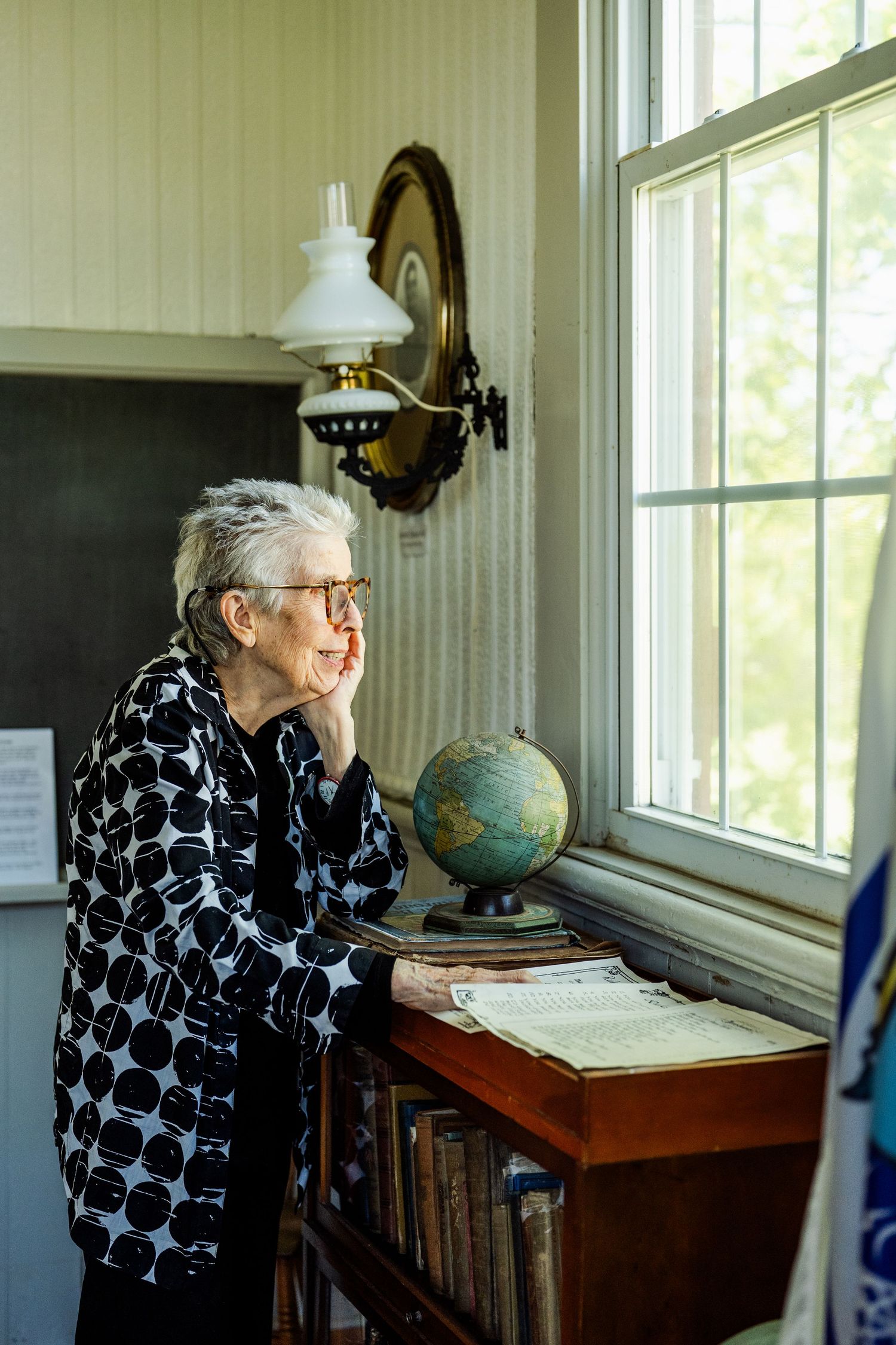 Person in black and white patterned shirt examines an antique desk with vintage globe and windows in background.