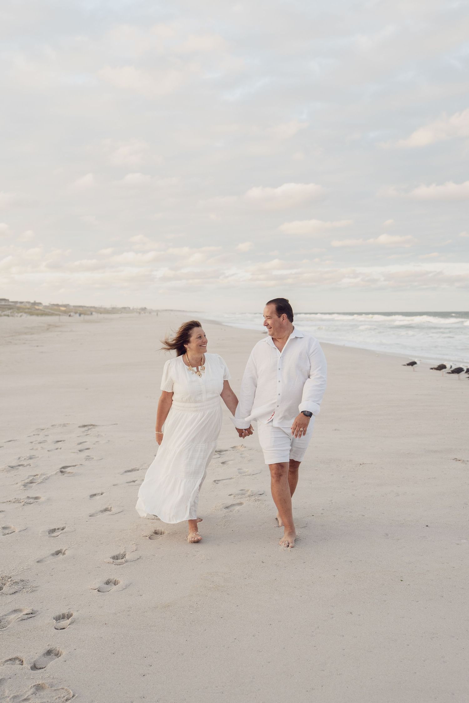 Couple strolling hand in hand along peaceful shoreline in matching white outfits.