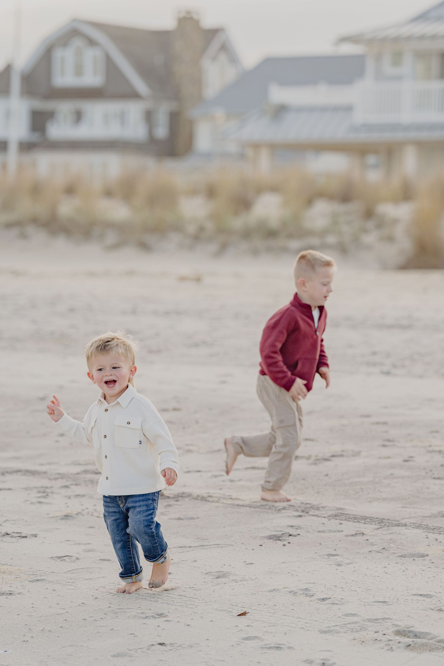 Two barefoot young boys run and laugh joyfully on a sandy beach with coastal homes visible in background.