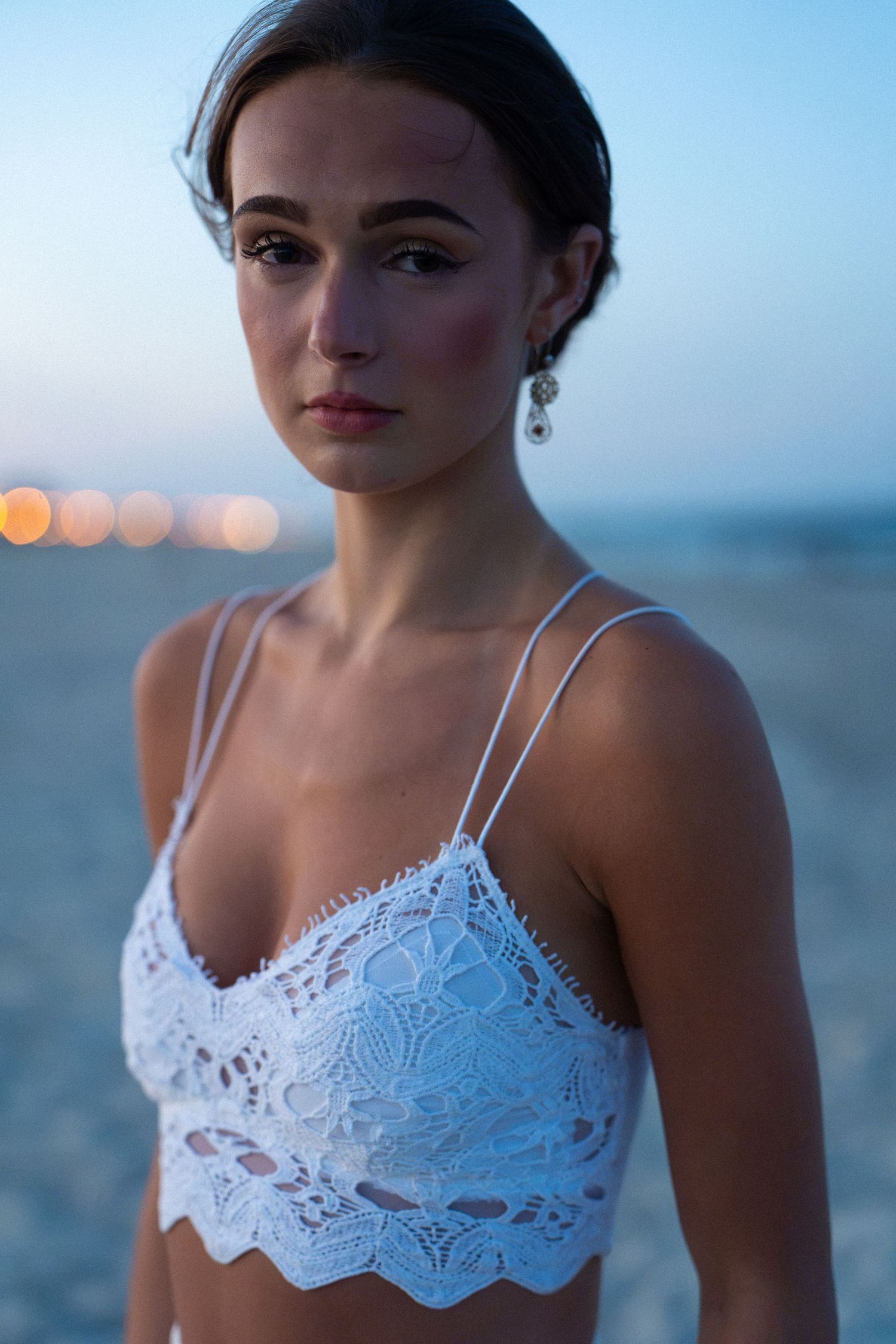 Young woman in white lace bralette with drop earrings poses on beach at dusk with bokeh lights behind.