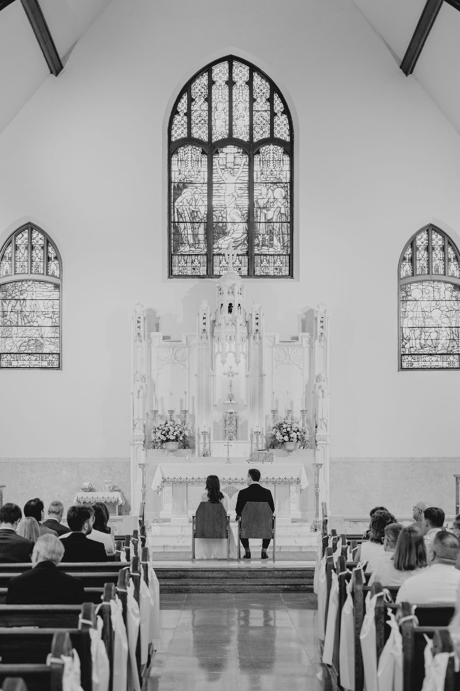 Bride and groom kneel at ornate church altar with stained glass windows during wedding ceremony.
