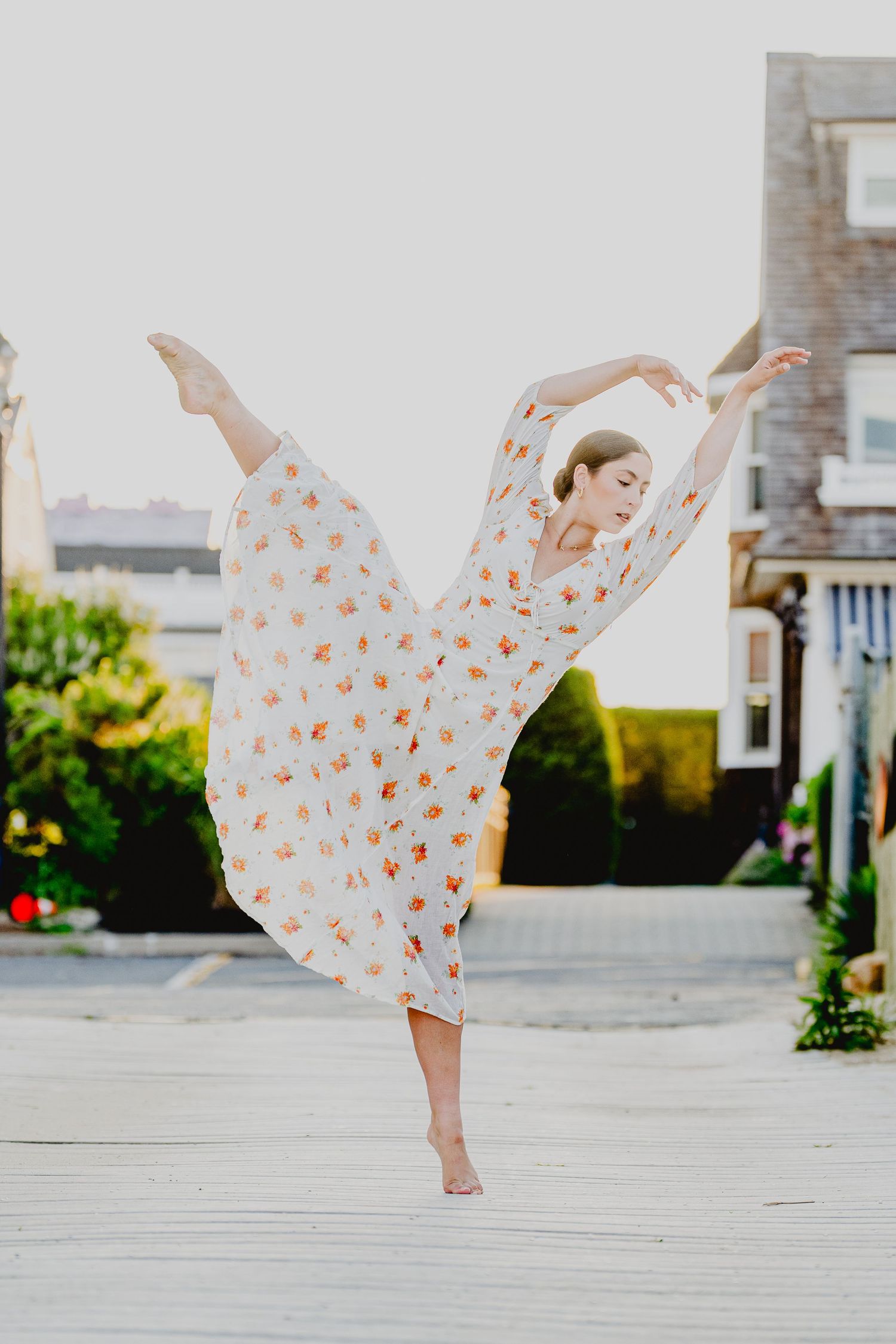 A graceful ballet dancer poses en pointe outdoors in a flowing white floral dress, arms arched overhead.