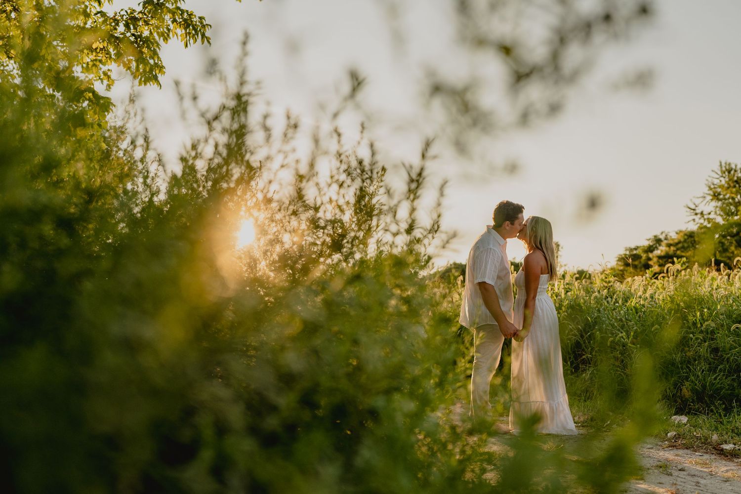 Couple kissing at golden hour surrounded by lush greenery, sunlight glowing behind them in a romantic outdoor setting.