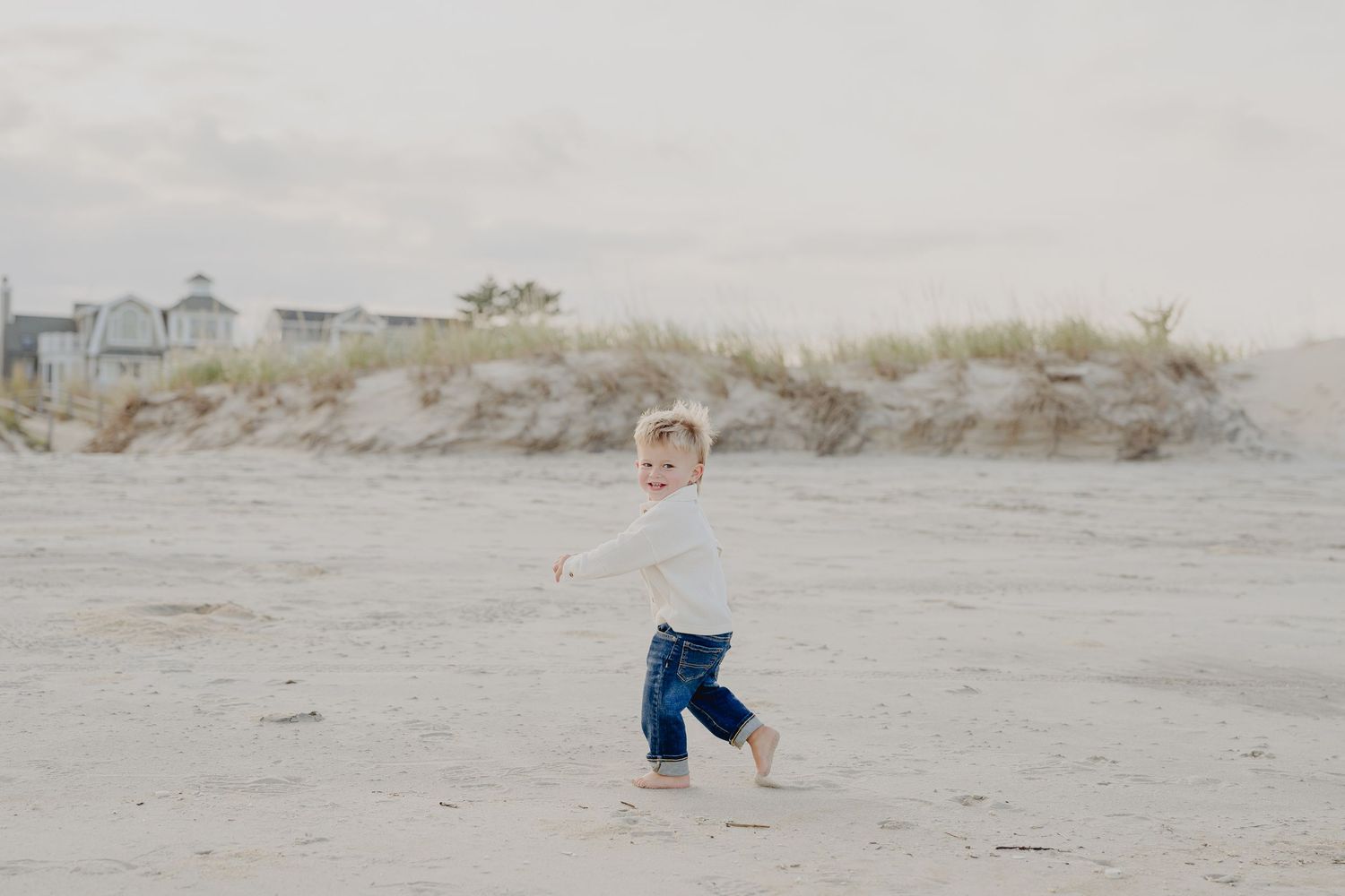 A toddler runs barefoot on a sandy beach wearing jeans and a white shirt with dunes and houses in the background.