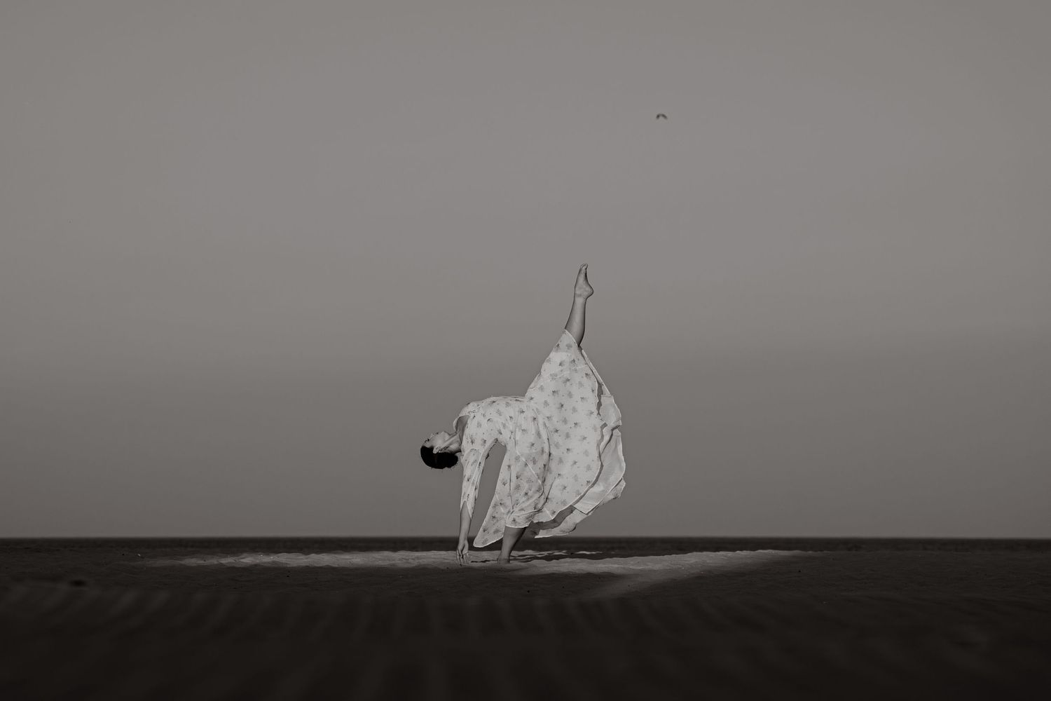 Black and white photo of a dancer in a flowing white dress bending gracefully on a sandy beach.