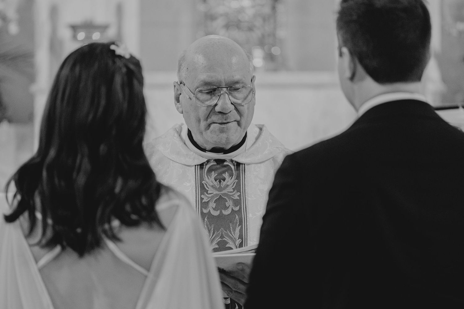Elderly priest officiating a wedding ceremony, facing the bride and groom in a church, black and white photo.
